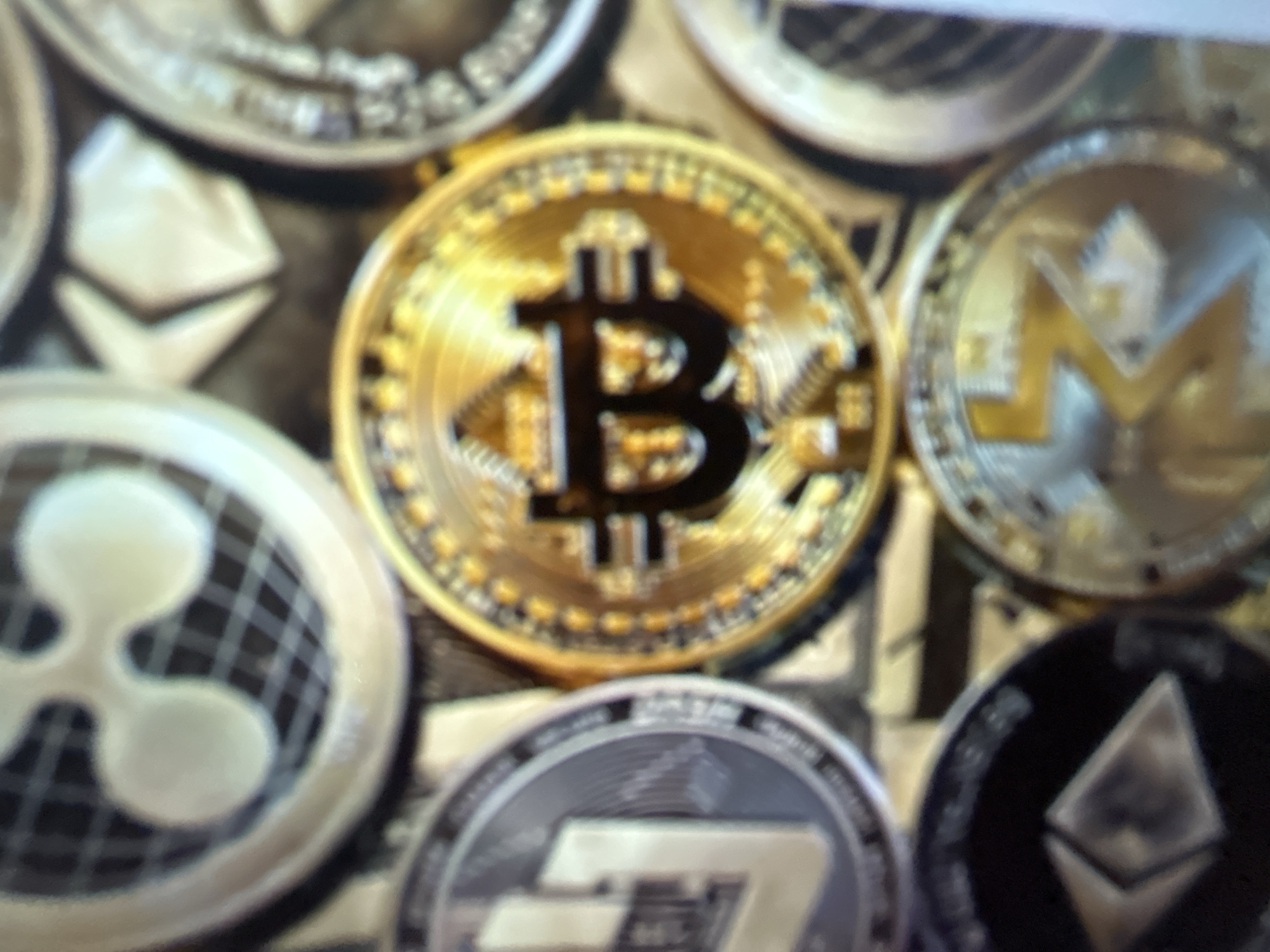 The image is a close-up, angled shot of several cryptocurrency coins, including Bitcoin, Litecoin, and Ripple. The coins are layered and out of focus, with the Bitcoin coin in the center being the most prominent and sharpest. The lighting is bright, highlighting the metallic sheen of the coins. There are no people or recognizable landmarks present, and no context is provided to suggest a location in Gelendzhik, Russia, or any specific city.