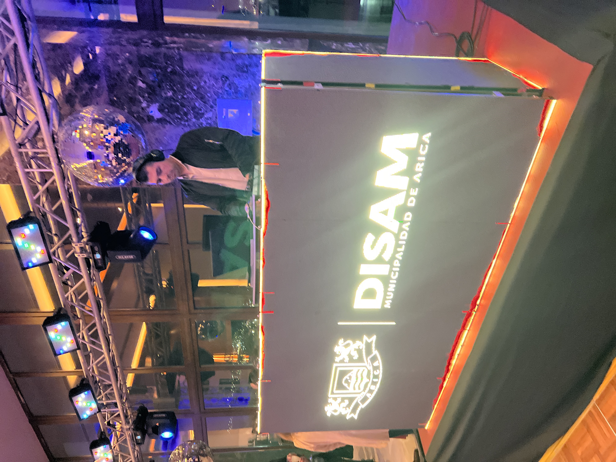 In an indoor venue, a male DJ wearing headphones and a black jacket over a light-colored shirt stands behind a large, illuminated DJ booth. The booth displays a logo with two lions flanking a crest that reads "ARICA," and the text "DISAM MUNICIPALIDAD DE ARICA" in large, bright letters. Above the DJ, a disco ball hangs from a truss system, which also supports multiple colorful stage lights. The background features large windows reflecting interior lights and a cityscape at night. The DJ is actively engaged with his equipment.