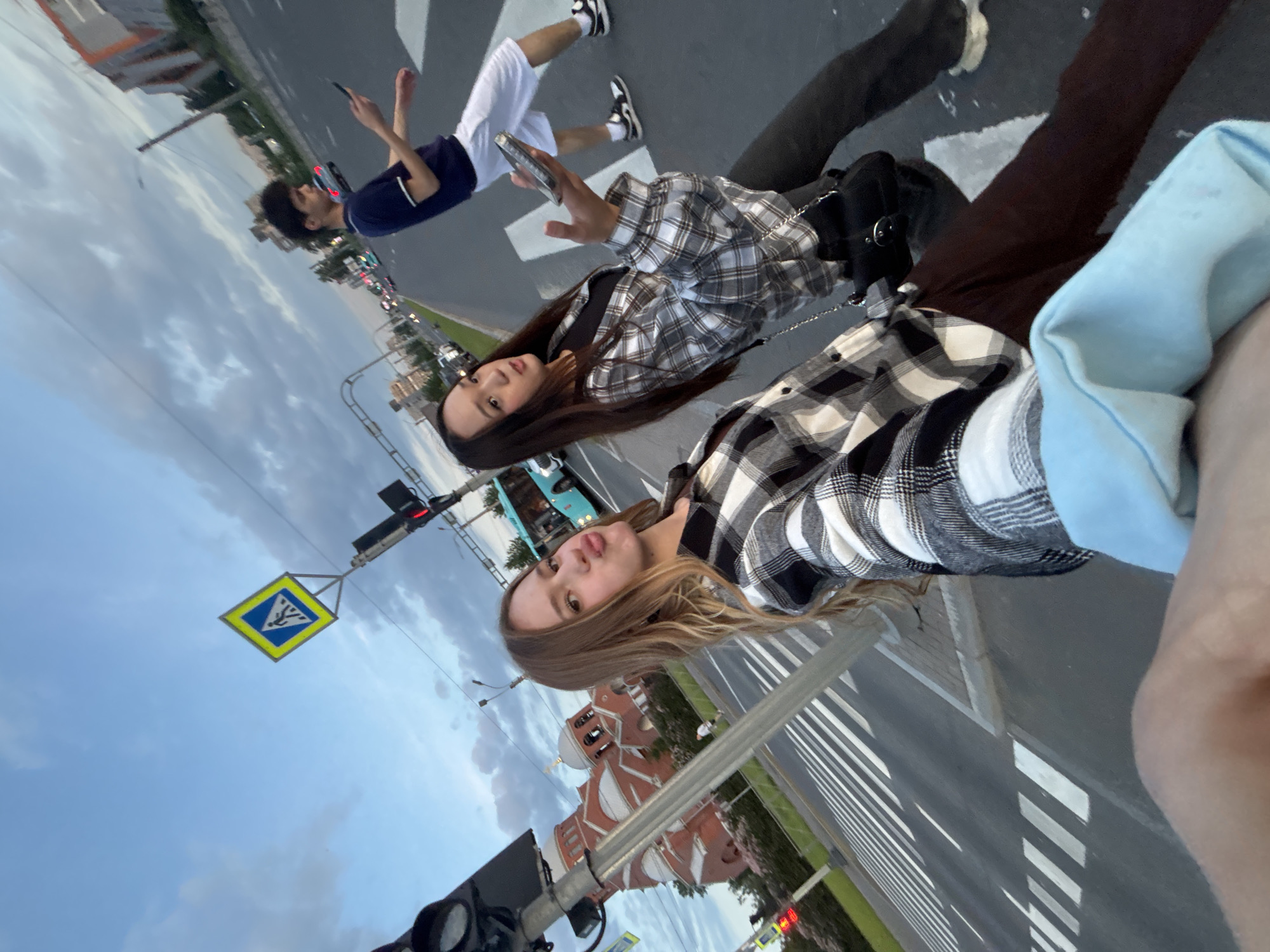 This is a selfie taken outdoors in an urban environment, likely in Russia due to the presence of a pedestrian crossing sign consistent with Russian road signage. Three young adults are visible, two women in the foreground and one male in the background. The two women are posing for the camera, with the woman on the left making a duck face. They are both wearing plaid shirts. The woman on the right is holding a smartphone. The man in the background is walking across a pedestrian crossing and looking at his phone. A red brick church with a dome is visible on the left side of the image. There is a bus in the background, and the sky is overcast. The scene appears to be a street with traffic lights and crosswalks.