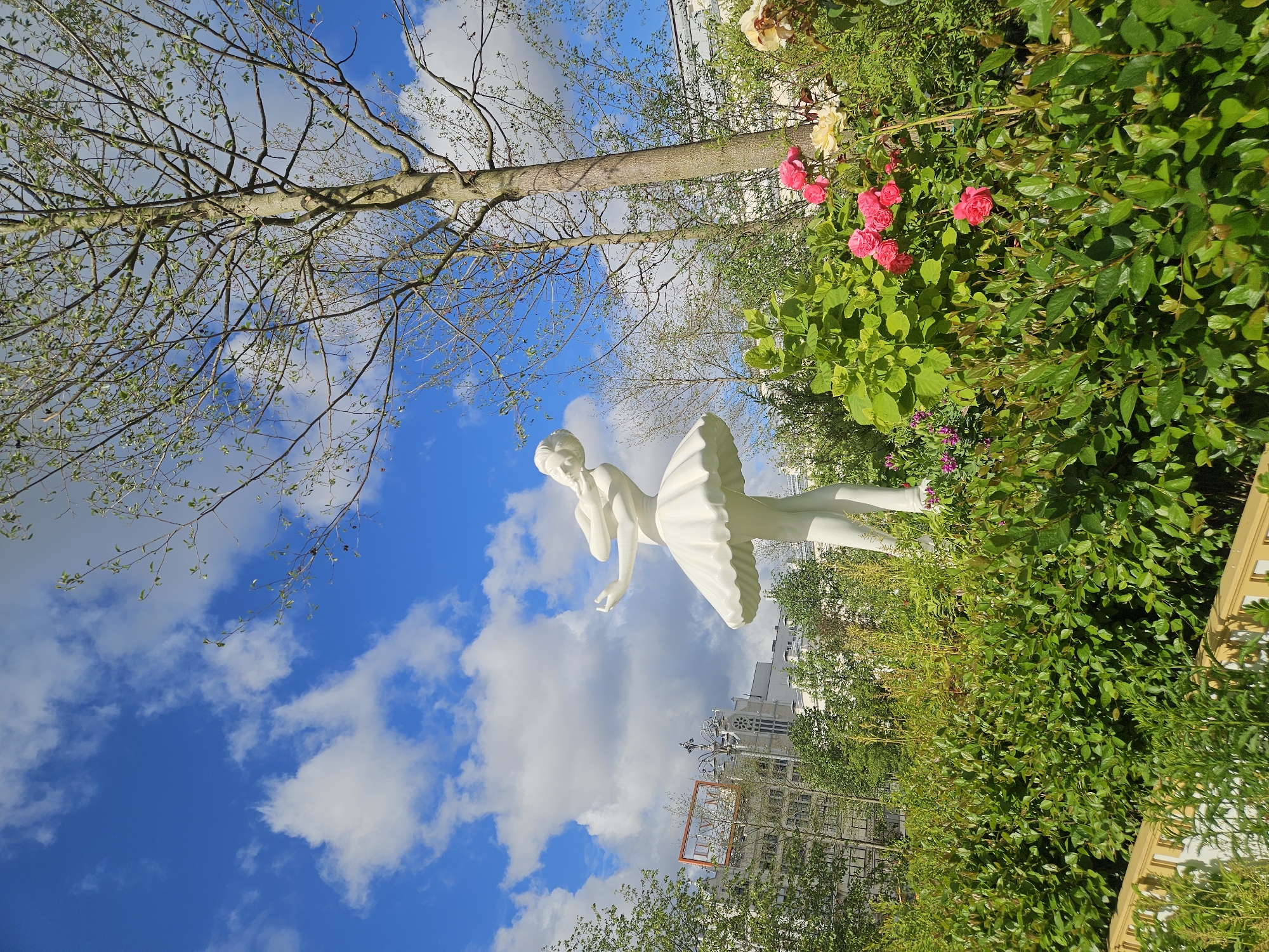 The image depicts a statue of a ballerina in a garden setting against a partly cloudy sky. The statue is white, with the ballerina in a classic pose, suggesting motion. The surrounding flora includes roses and dense green foliage. A tree branch extends from the top left of the frame across the sky, indicating a natural setting within the urban landscape. A building and what appears to be a sign in Cyrillic characters can be seen in the background. The location appears to be near the GUM department store in Moscow, Russia.