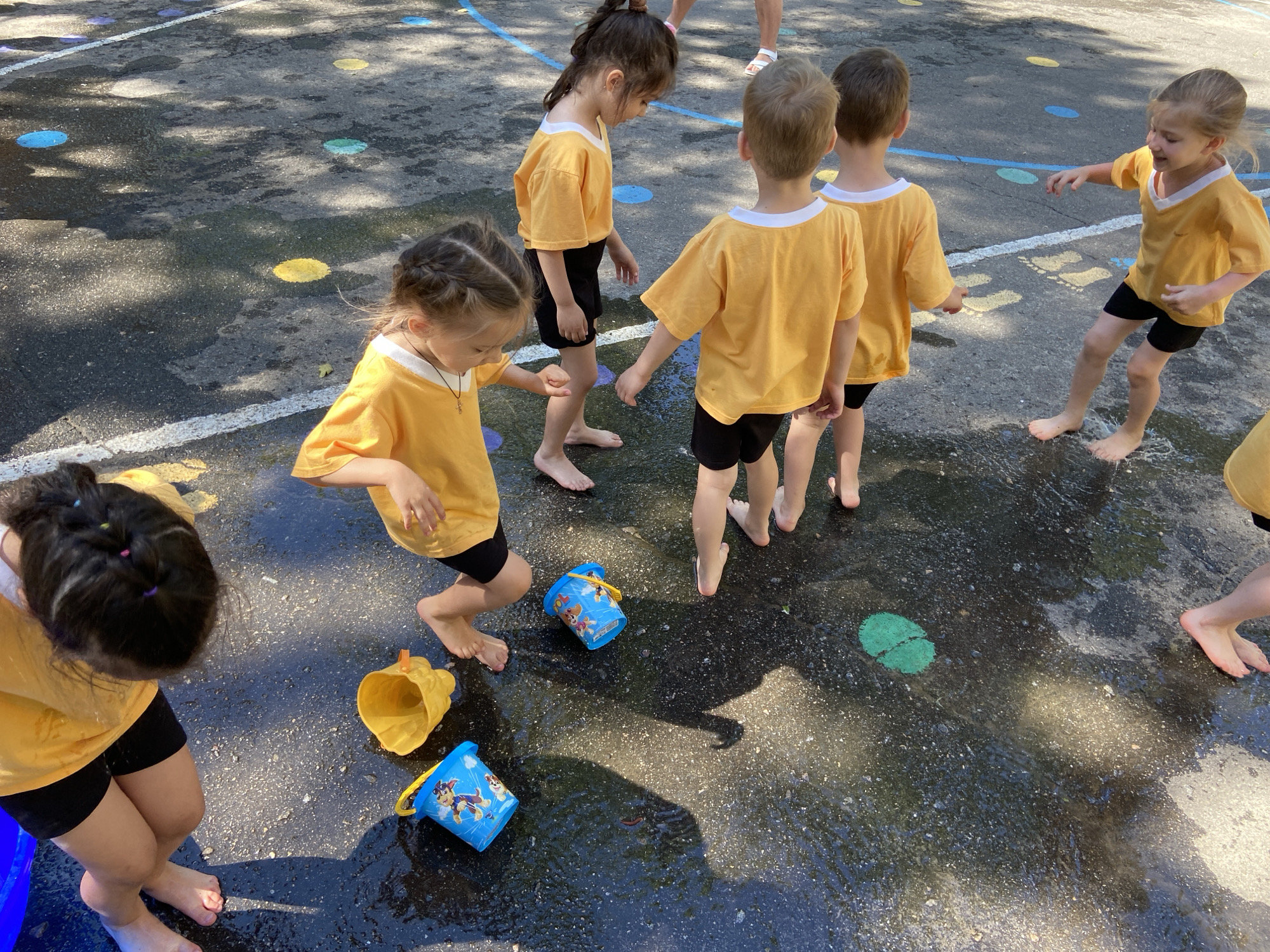 The image shows a group of young children, likely toddlers or preschoolers, playing in a wet, outdoor play area. The children are wearing matching yellow t-shirts and black shorts, and are all barefoot. The ground is asphalt with colorful dots (blue, yellow, green) painted on it, and some areas are wet, indicating water play.
Several blue buckets, one with a Paw Patrol design, and a yellow funnel-shaped toy are visible on the ground amidst the puddles. The children appear to be engaged in some sort of water activity, possibly a splash pad or a water game. One girl in the foreground is actively splashing water with her foot, her face showing excitement. Another girl to her left is looking down at the ground, her hand near a bucket. In the background, other children are also standing or moving around in the wet area.
The sunlight filtering through trees casts shadows on the ground, suggesting it is daytime, likely a sunny day. The overall mood seems joyful and energetic, as children are engrossed in play. The setting appears to be a playground or a designated outdoor play space. No visible text is present in the image itself, other than the design on the bucket.