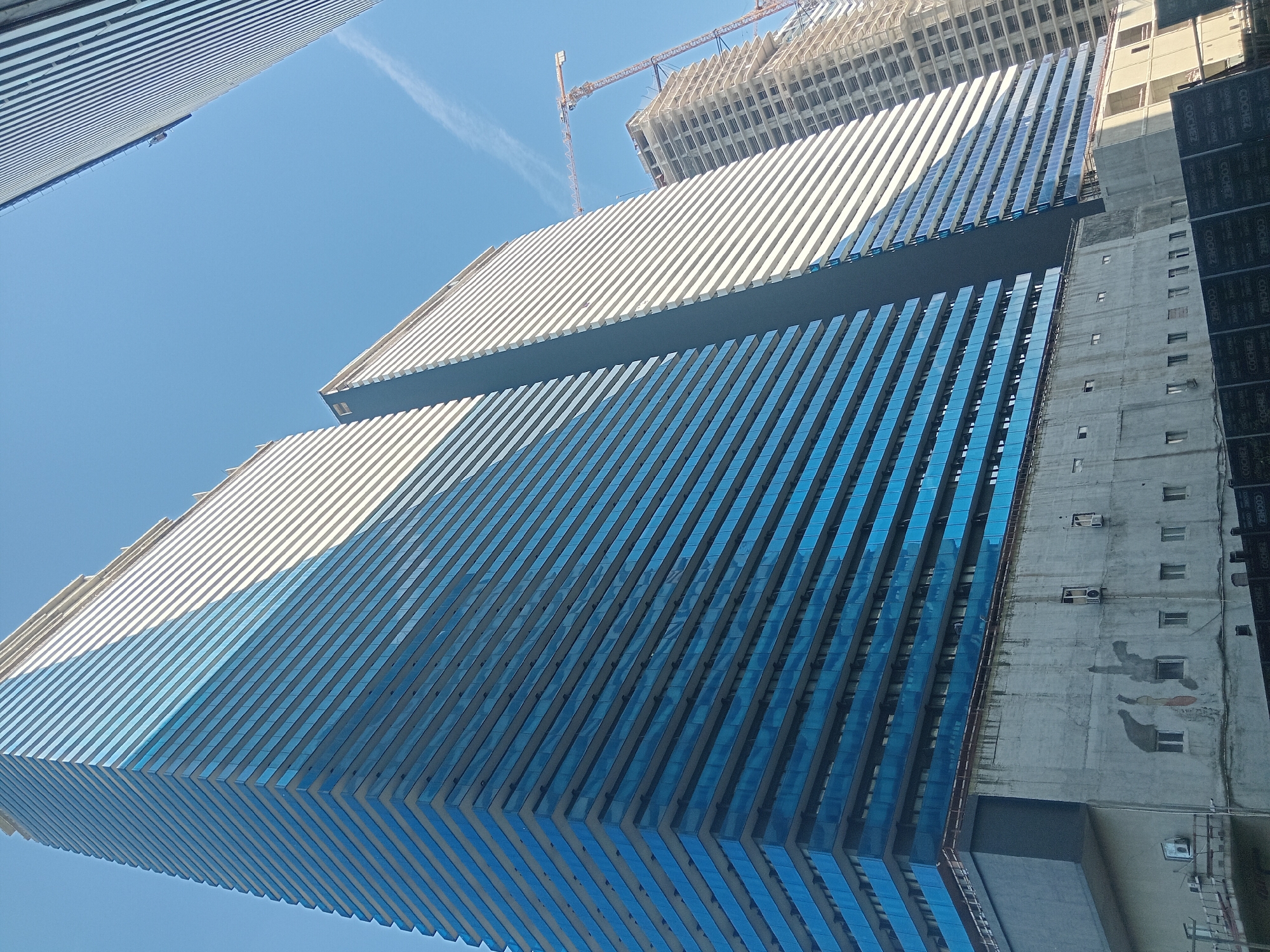 The image captures a low-angle perspective of modern buildings in Batumi, Georgia. Two high-rise buildings dominate the frame, featuring a design with horizontally oriented elements, predominantly in shades of white and blue, suggesting glass and metal construction. The buildings exhibit a repetitive, lined pattern.
A crane is visible in the background near another building under construction, indicating ongoing development in the area. The sky is clear and bright blue. A neighboring building of bare concrete, partially covered with black panels and utility installations like AC units, is adjacent to one of the glass buildings. The overall scene indicates a modern urban environment with active construction sites.