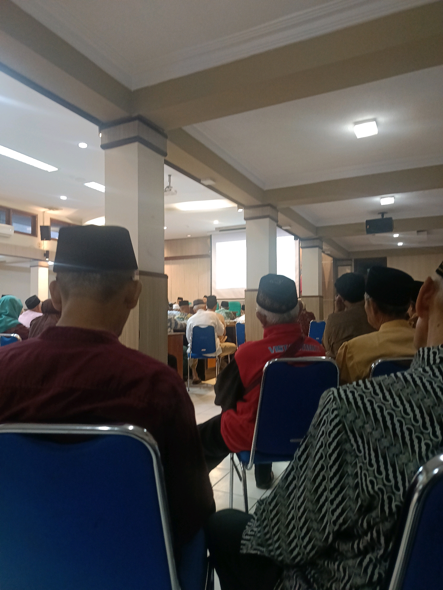 The image shows an indoor gathering of people, likely a meeting or presentation, in a room with fluorescent lighting and beige ceiling beams. Several individuals are seated, facing away from the camera, many wearing traditional Muslim attire including peci (caps) and modest clothing. In the foreground, a person wearing a maroon shirt and a black peci is prominently visible from behind. Other individuals are seated in rows of blue chairs. In the background, a projector screen displays a presentation, and there are several pillars supporting the ceiling. The overall impression is that of a formal or semi-formal event taking place in a community hall or meeting room. The image is likely taken in the city of Yogyakarta, Indonesia, given the cultural context suggested by the attire and the commonality of such events in the region.