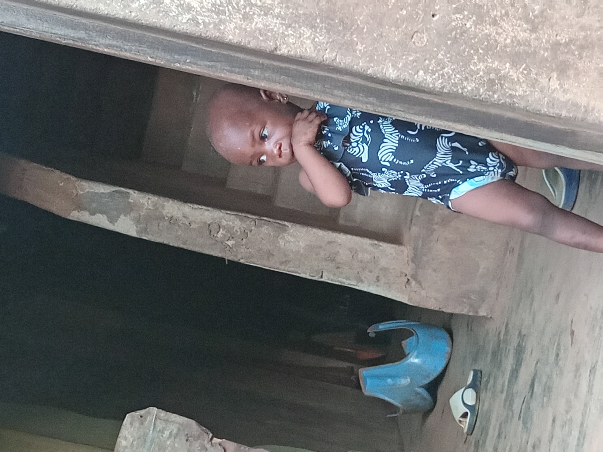A young child, appearing to be a toddler, peeks curiously from a shaded, rustic doorway or passage. The image is oriented diagonally, with the child's head tilted towards the left. The child has dark skin and a bald head, with a small, light-colored earring visible in their left ear. They are dressed in a dark blue romper or bodysuit patterned with numerous white, stylized animal figures, possibly tigers or zebras. One of the child's hands is raised and placed near their mouth or chin, giving them a thoughtful, shy, or slightly pensive expression as they look directly towards the viewer.
The setting is characterized by rough, textured walls and a ground made of concrete or dried earth, indicative of a simple, unadorned environment in Shagamu, Nigeria. The area is dimly lit, suggesting shade from the daytime sun. On the dusty floor beneath the child, a distinctive bright blue plastic object, possibly part of a toy or a container, is visible. Nearby, a single white and black sandal or slipper lies on its side. No specific activity is taking place other than the child observing the scene. There is no visible text in the image.