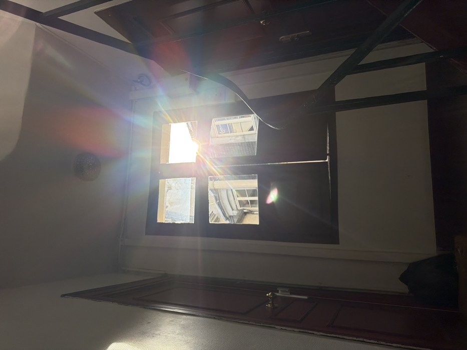 This image depicts a static interior view of a sunlit stairwell or landing within a building in Paris, France. A central, dark-framed window with four panes allows strong natural light to penetrate, causing prominent, multi-colored lens flares across the upper half of the image, emanating from the upper-right window pane. Reflections in the window reveal typical Parisian architecture, specifically in the lower-left pane, which shows a light-colored building facade with a distinct curved bay window and a visible internal window.
On the left, a dark red/brown paneled door with a brass-colored doorknob and lock plate stands against a light-colored, textured wall. On the right, a similar dark red/brown paneled door is partially obscured by a dark, possibly black, metal staircase handrail, which curves upwards, indicating an adjacent staircase. A round, textured silver-colored light fixture is mounted on the light-colored ceiling. The walls are generally light-toned, with sections illuminated by direct sunlight and others in shadow. A dark, indistinct object, possibly a bag, rests on the dark floor near the bottom of the left door frame. No individuals are present, and no actions or interactions are depicted.