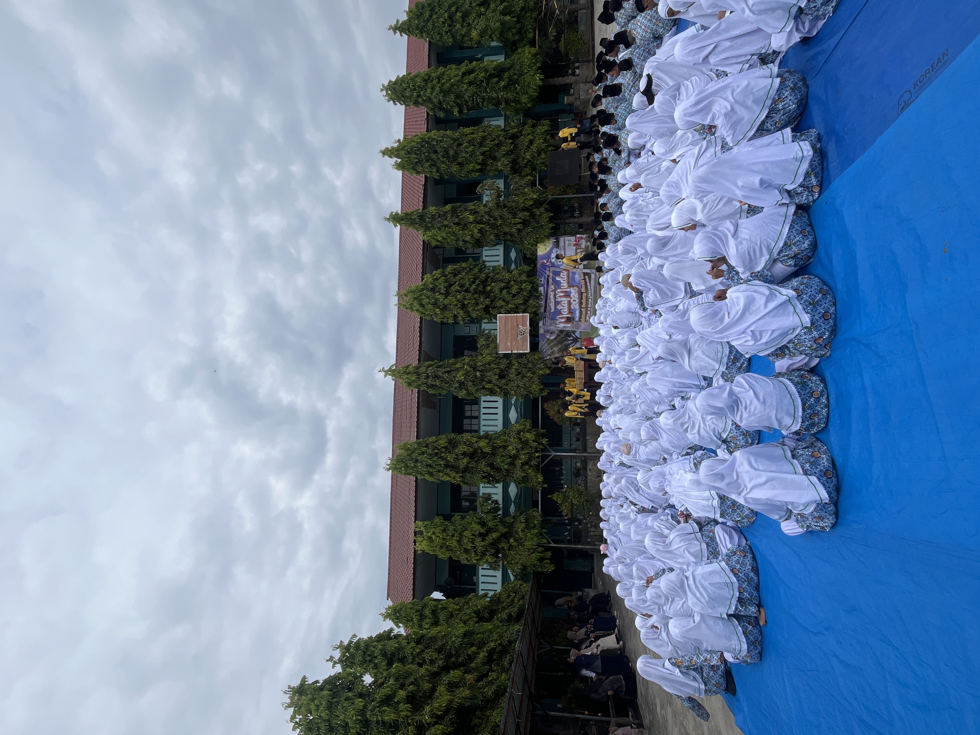 The image depicts an outdoor gathering of many individuals, predominantly young women wearing white hijabs and patterned skirts, seated on a large blue tarp. They are facing a stage area with a backdrop featuring text that appears to be an event banner. Behind the audience and stage, a building with a red roof and green facade is visible, flanked by tall, slender trees. Several people, some wearing yellow shirts, are on or near the stage. The sky is overcast with gray clouds. The scene suggests an event taking place in an educational institution or public space in Lhokseumawe, Indonesia.