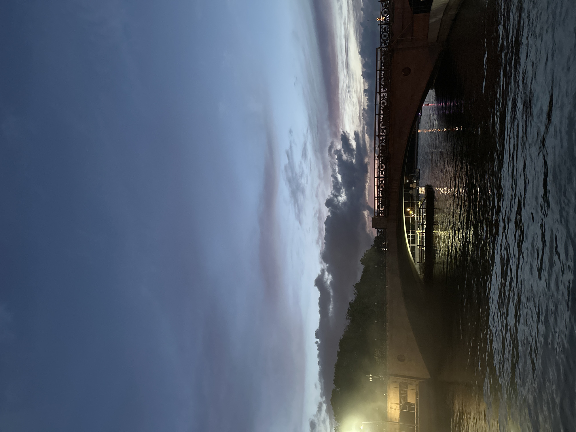 A dusky sky with streaks of pink and purple clouds hangs above a body of water. A stone bridge arches over the water, with lights from a boat docked beneath it casting reflections on the surface. The bridge is adorned with an ornamental railing, and trees line the bank behind it. Streetlights illuminate the walkway on the bridge and the bank. The overall scene is one of twilight in an urban setting.