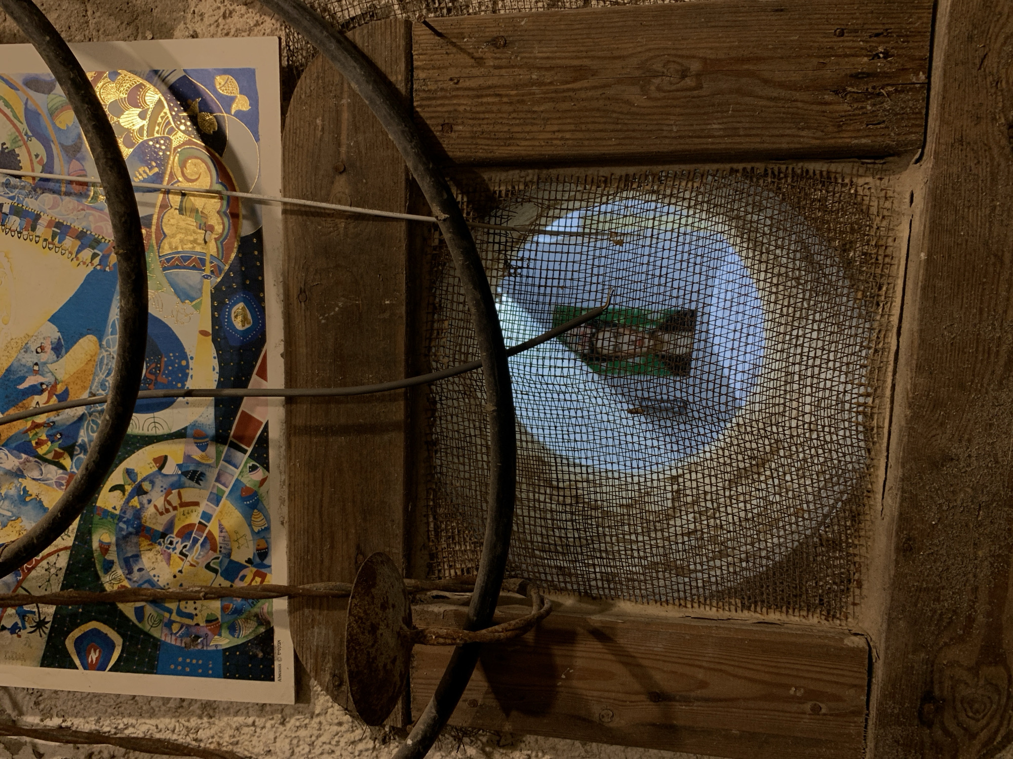 The image shows a close-up view of a section of a wall, possibly indoors. There's a framed window covered with a wire mesh screen. The window is set into a wooden frame. A colorful poster is partially visible, leaning against the wall to the left of the window. A thick black cable runs across the scene. The poster contains designs of geometric forms and symbols. The general aesthetic suggests an old or rustic setting. The location in Tel-Aviv, Israel, is not discernible from the image.