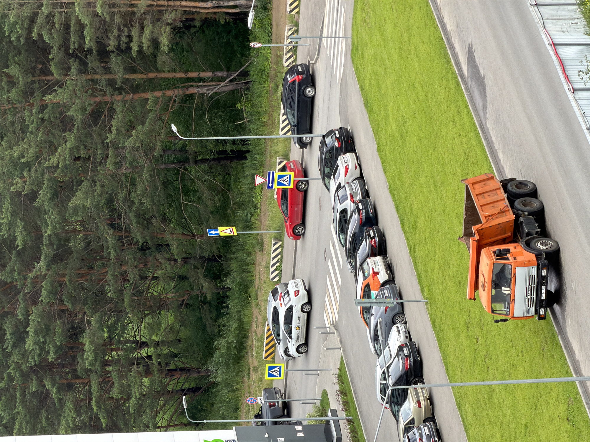 An aerial view captures a street scene in an urban environment, characterized by a dense forest of pine trees bordering a paved area with a parking lot and a road. In the foreground, an orange dump truck is parked on the side of the road, facing right. The truck has the brand name "KAMAZ" visible on its grille. To the left of the truck, a grassy embankment slopes upwards. Further back, a crosswalk is marked on the road, with several cars parked in a lot adjacent to it. A white taxi with "Яндекс Такси" branding is visible at the crosswalk, along with other parked vehicles including a silver sedan, a dark sedan, and an orange hatchback. A traffic light pole with signage is present near the crosswalk. In the background, more cars are parked, and the dense pine forest forms a significant portion of the scene, with the trees extending to the horizon. The sky is not visible.