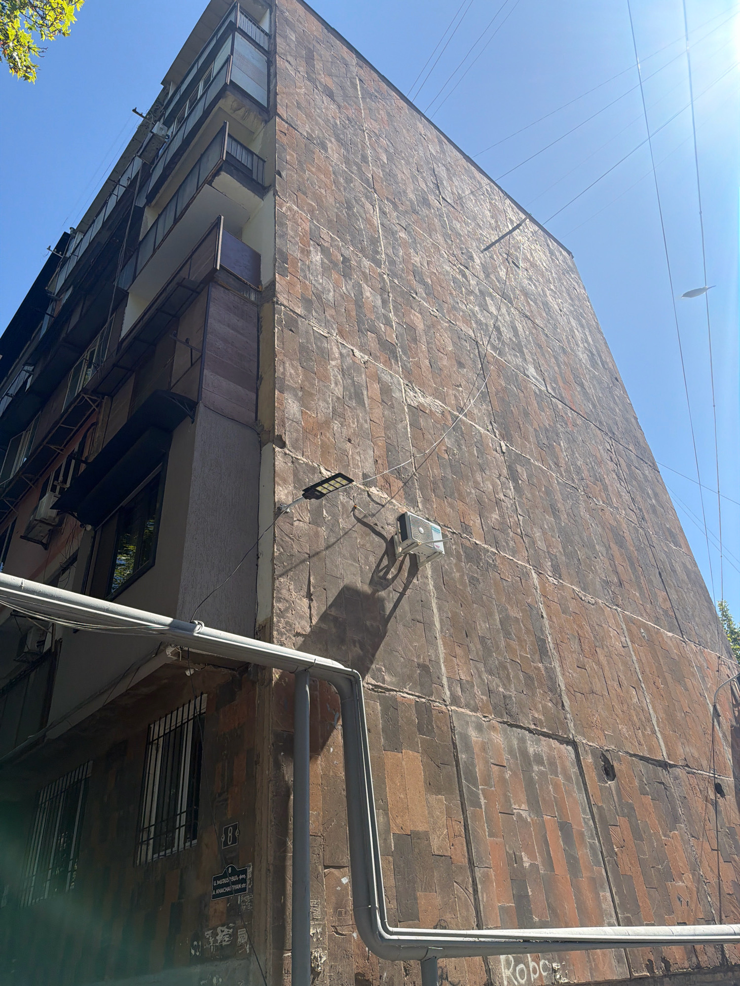 The image depicts the exterior of a multi-story building in Yerevan, Armenia, characterized by a facade of large, reddish-brown stone tiles. The building appears to be an apartment complex, with balconies visible on the left side. Several air conditioning units are mounted on the exterior walls. Electrical wires and a street lamp are also present. The lower section of the building shows some signage, including what appears to be an address or building number. The scene is captured on a clear, sunny day, with a blue sky visible above. The perspective is from a low angle, looking upwards at the building.