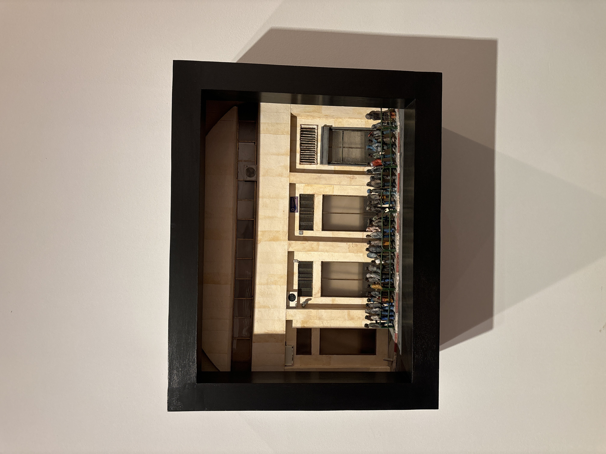 The image depicts a framed three-dimensional artwork hanging on a white wall. The artwork portrays a building facade, likely from Tel-Aviv, Israel, characterized by its light-colored stone construction and architectural style typical of the region. The facade features windows with security bars, air conditioning units, and a narrow balcony or ledge with an array of objects, possibly shoes or other personal items, arranged along it. The building's architecture is relatively simple, with vertical and horizontal lines defining its structure. The artwork is presented in a black, deep-set frame. Shadows from the artwork and frame are cast onto the white wall, indicating a light source positioned above and to the right of the artwork. The environment is stark and minimalist, emphasizing the artwork as the primary subject.