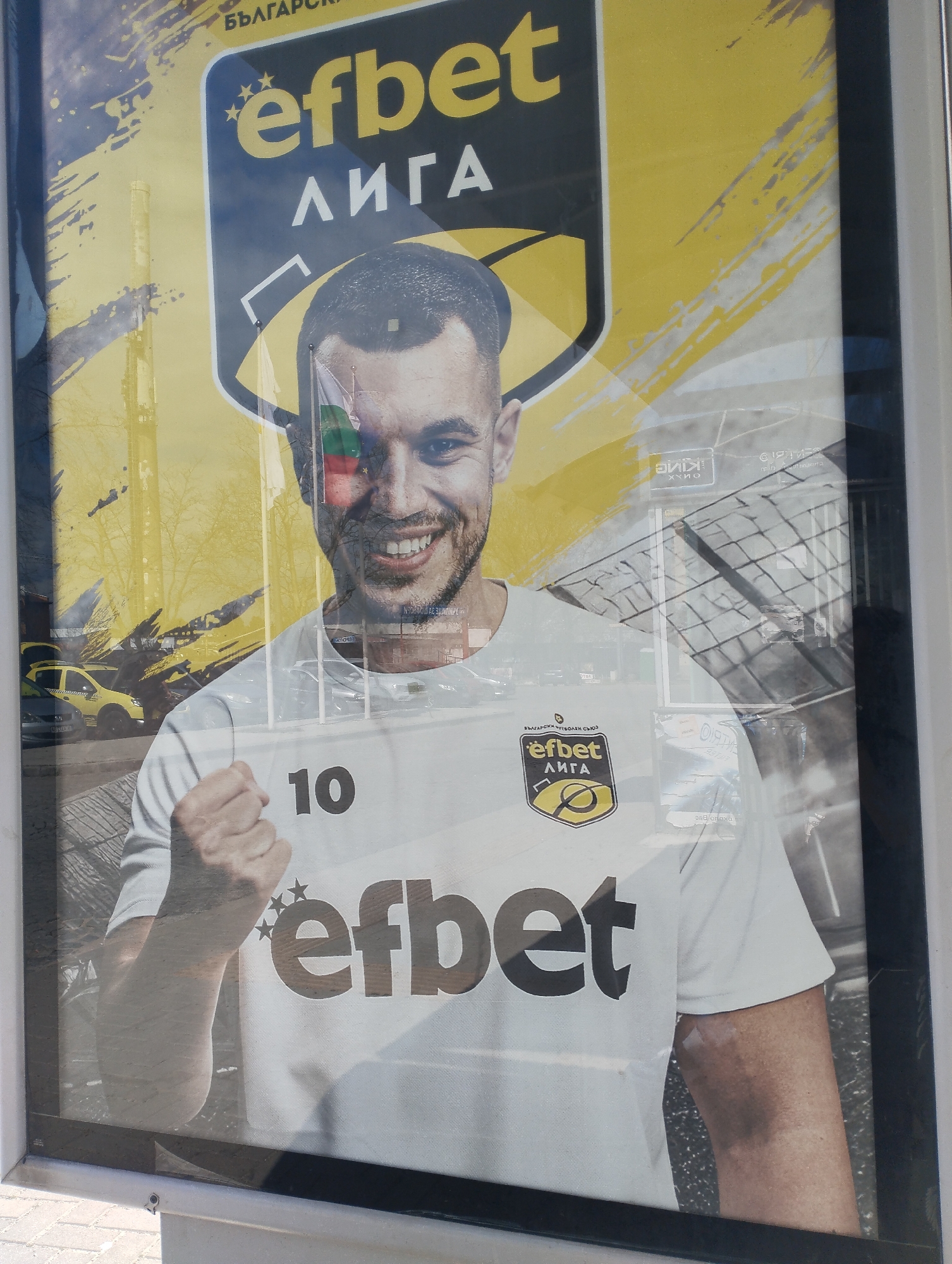A close-up photograph shows a poster advertising "efbet лига," a Bulgarian football league. A smiling man, possibly a football player, wears a white jersey with the number "10" on the shoulder and the "efbet" logo on the chest. The Bulgarian flag is positioned above the man's left eye. The poster is framed and appears to be displayed outdoors, reflecting a building, vehicles, and trees, suggesting the image was taken in the city of Yambol, Bulgaria.