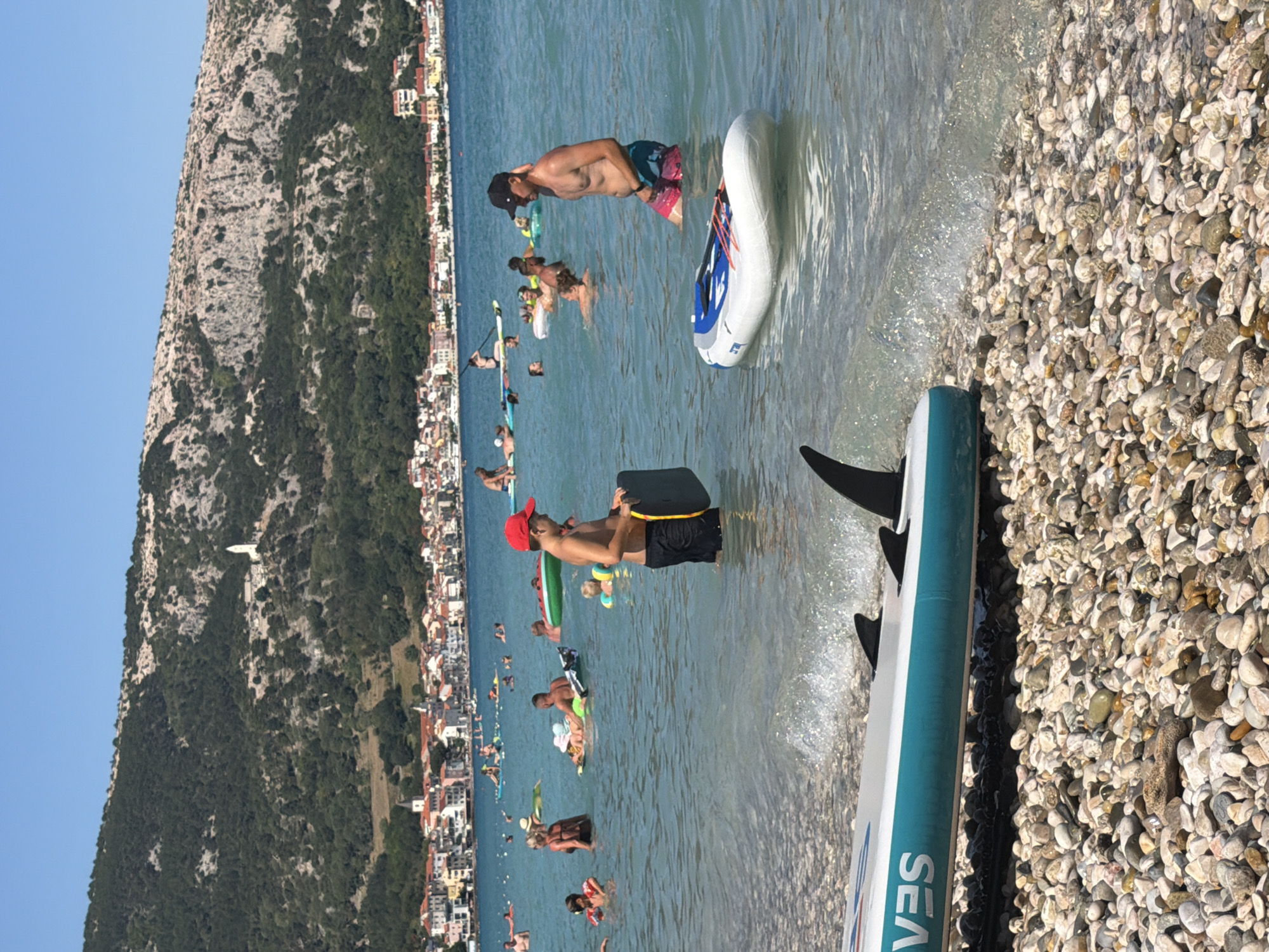 The image captures a vibrant scene at a beach in Baška, Croatia. The primary subjects are people enjoying a sunny day at the water's edge and in the shallow sea. Several individuals are visible, some standing in the water, others on paddleboards or inflatable rafts.
One man, wearing a red cap and black shorts, is holding a black bodyboard in the water. Another man, in patterned shorts and a black baseball cap, is standing in the sea, his arm behind his head. There are various inflatable objects in the water, including a large white and blue inflatable and several smaller, colorful ones. A paddleboard with a black fin is prominently positioned in the foreground, resting partially on the pebbled shore and partially in the water.
The setting is a beach characterized by a multitude of smooth pebbles lining the shore. The sea is clear and turquoise, with gentle waves lapping at the beach. In the background, a lush, green mountainside rises dramatically, dotted with buildings of a town nestled along the coastline. The architecture suggests a Mediterranean or Adriatic setting. The bright sunlight and clear blue sky indicate fair weather and likely a daytime scene, possibly late morning or afternoon given the strong illumination. The atmosphere appears relaxed and recreational, with people engaging in water activities typical of a summer beach day.
The visible text on the paddleboard in the foreground reads "SEA". The overall impression is one of a lively summer vacation spot, with natural beauty and recreational pursuits coexisting.