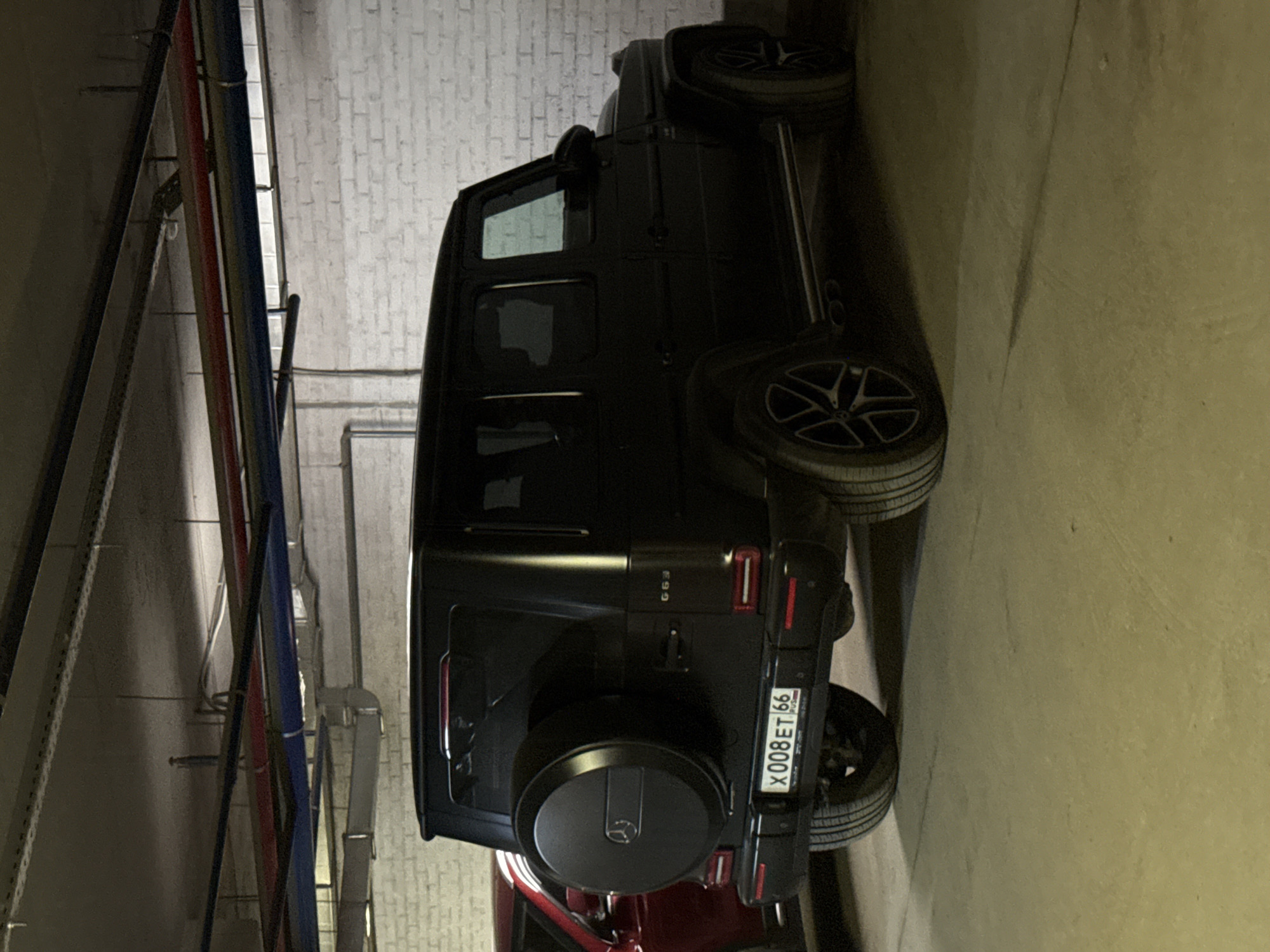 A vertically oriented image captures a black Mercedes-Benz G-Wagen, specifically a G63 model, parked indoors. The vehicle is viewed from its rear-left side, showcasing its rugged, boxy silhouette, dark-tinted windows, and multi-spoke black wheels. A spare tire, enclosed in a black cover featuring the Mercedes-Benz logo, is mounted on the rear tailgate. The "G63" emblem is visible on the tailgate.
The setting appears to be an underground parking garage or a basement area. The background features a textured white brick wall, while the foreground and right side are dominated by an off-white or beige concrete floor/wall. Overhead, an intricate network of utility pipes, cables, and electrical conduits, in colors such as red, blue, grey, and white, runs along the ceiling, characteristic of industrial or building infrastructure. The lighting is artificial and dim, typical of such indoor environments.
Part of a red car is visible parked immediately in front of the black G-Wagen. No individuals are present in the image.
A key detail is the Russian license plate clearly visible on the G-Wagen's rear: "X008ET 66". The "66" region code identifies the vehicle as registered in Sverdlovsk Oblast, which includes the city of Yekaterinburg, aligning with the provided location context. The license plate also displays the "RUS" country code and a small Russian flag. The scene is static, capturing vehicles at rest in an indoor facility.