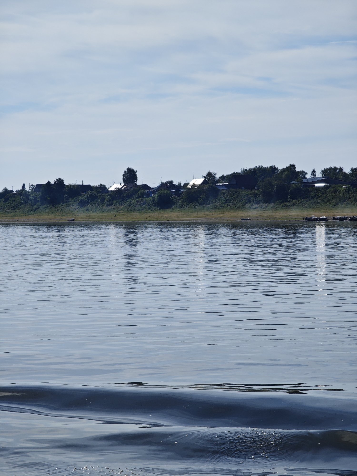 The image depicts a wide, calm river with a shoreline in the background. The river occupies the majority of the foreground and midground, with subtle ripples and reflections from the sky and the distant shore. On the far bank, a line of trees and vegetation forms a dense band across the horizon. Dotted within this greenery are several buildings, appearing to be houses or other rural structures, with visible roofs. Some smoke or mist seems to be rising from the ground in front of the buildings. A few boats are visible pulled up onto the riverbank. The sky above is a pale blue with scattered white clouds. The scene conveys a sense of a tranquil, perhaps rural, setting on a clear day. It is difficult to definitively identify the location as Yartsevo, Russia, without further contextual information or landmarks.