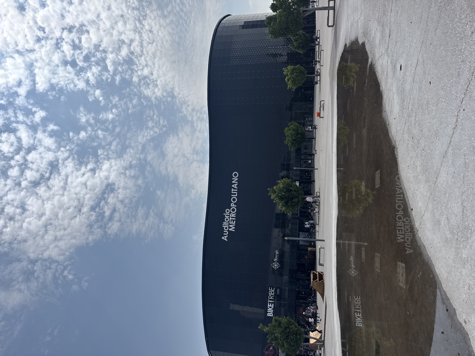 A ground-level, outdoor shot, rotated counter-clockwise, presents the Auditorio Metropolitano in Puebla, Mexico. The dark, curved facade of the building prominently displays the name "Auditorio METROPOLITANO" in large, white letters. Logos of "BIKETRIBE" and "Sinenergia" are also visible. A row of trees lines the side of the building, and multiple parked bicycles suggest activity in the vicinity. The clear sky features many white cumulus clouds. A large puddle on the pavement reflects the building and its surrounding elements, including the logos and trees. A few unidentified people are visible on the sidewalk. The pavement itself appears to be textured concrete.