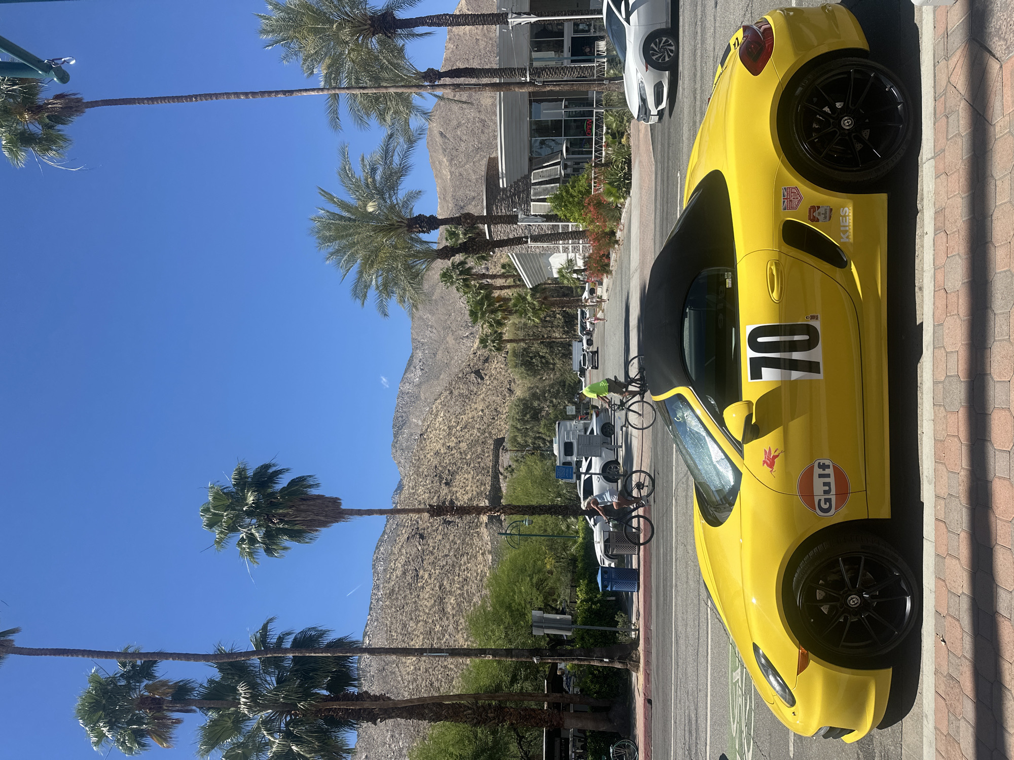 A yellow convertible sports car with racing decals and the number "70" on the side is parked on a brick-paved street, its passenger side facing the camera. The car is parked next to a road with bicycle lanes. Behind the car, palm trees line the street, with a mountain range in the background. A few vehicles and people on bicycles are visible further down the street. Buildings are also visible in the background. The sky is clear and blue. The location appears to be in a city but the specific city is unknown.