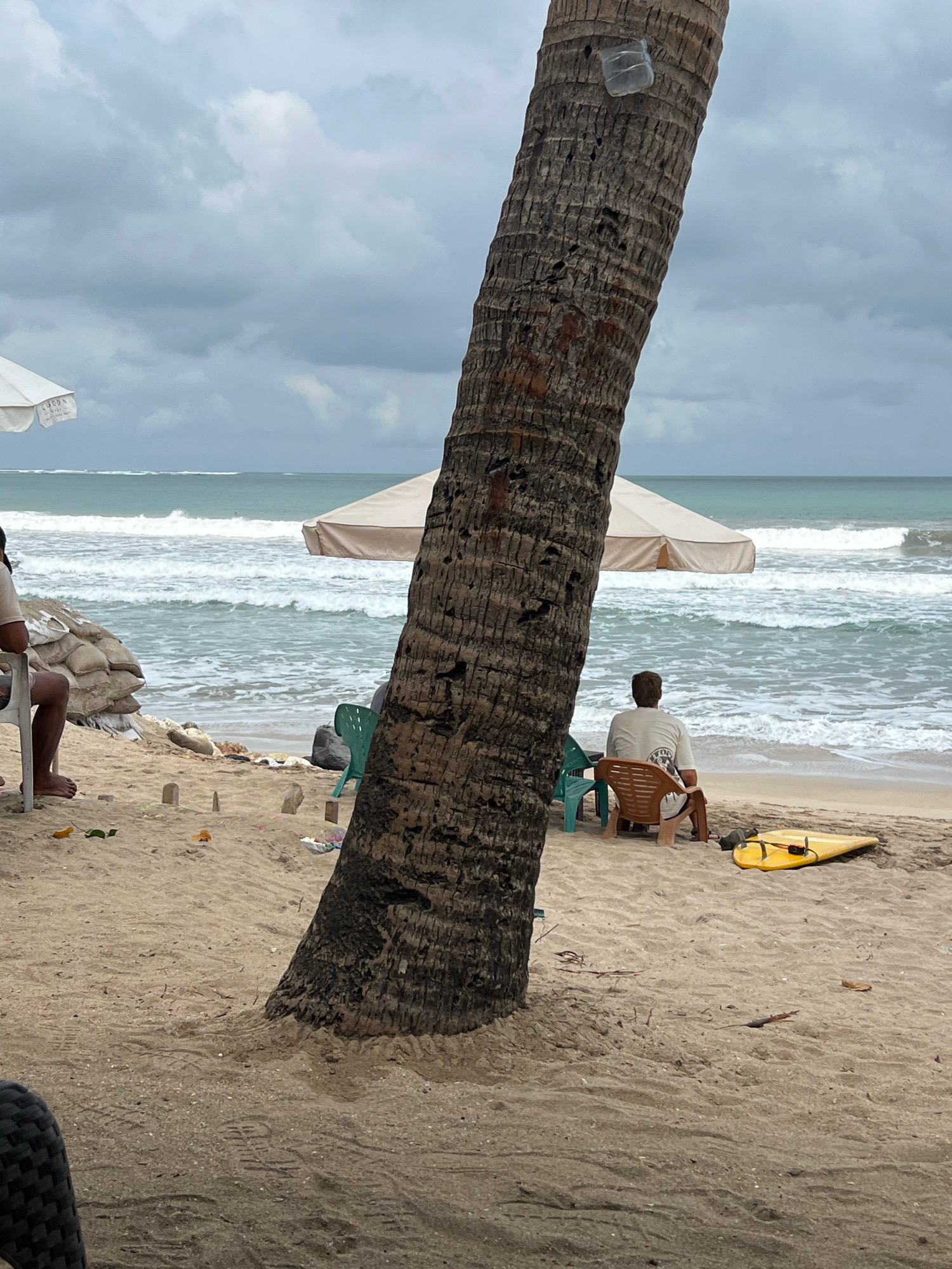 In Kuta, Indonesia, on a sandy beach under a cloudy sky, a large palm tree trunk dominates the foreground. Behind it, the ocean displays moderate waves. To the left, a white umbrella is visible, with a person sitting on a white plastic chair, their legs and feet bare. Nearby are sandbags. Further to the right, a person sits in a brown plastic chair facing the ocean. A small yellow surfboard lies on the sand near them. Two green plastic chairs are also present. The beach is strewn with debris, including leaves and small wooden markers.