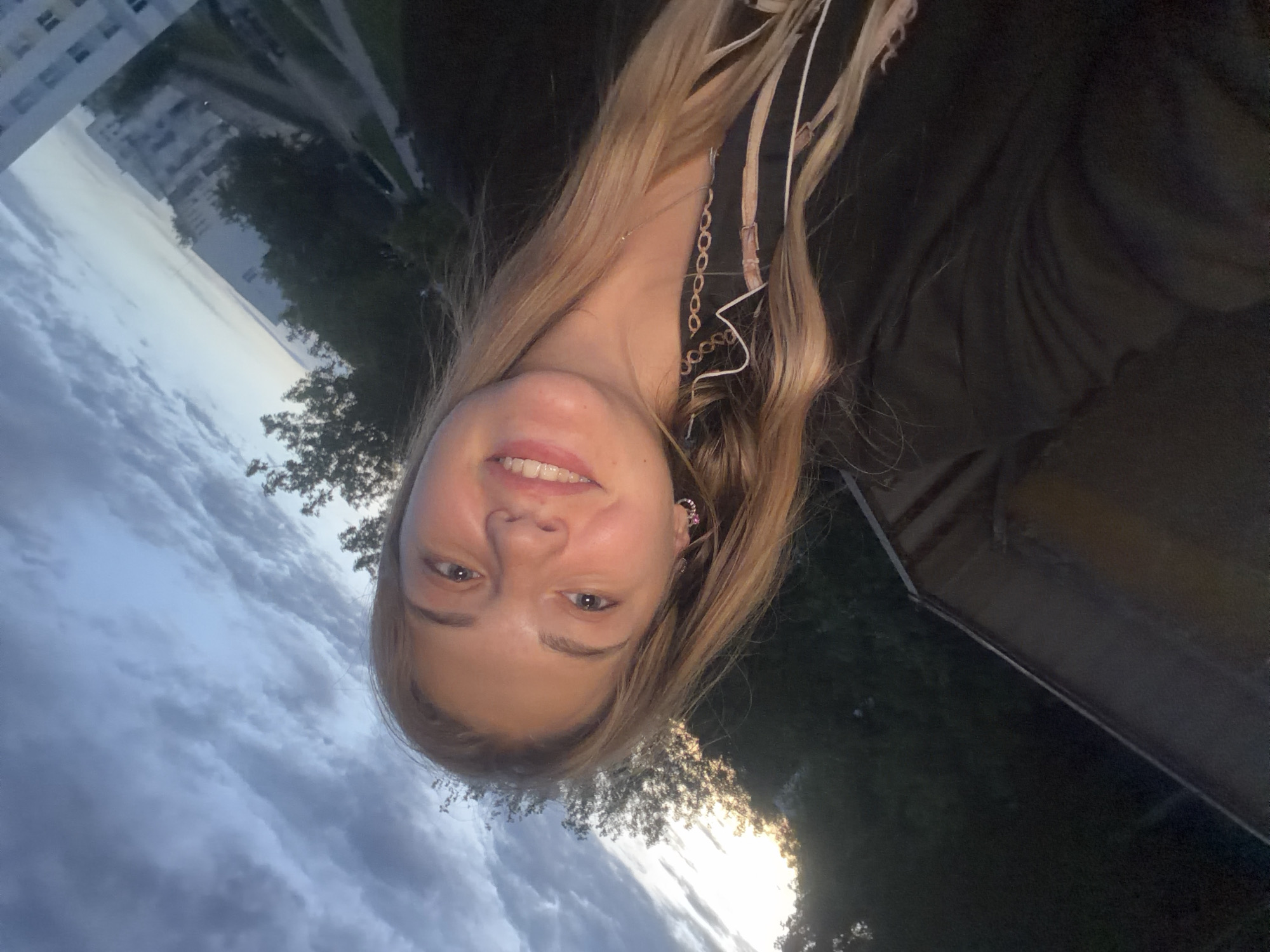 A medium shot of a young woman with long blonde hair, facing forward and smiling. The background shows a cloudy sky and foliage. In the distance, a white, multi-story building is visible, likely a residential apartment block. The setting appears to be an outdoor area, possibly a park or residential zone. The presence of a modern building and dense vegetation suggests the location may be in the city of Naberezhnye Chelny, Russia. A white object with a cord is hanging from her neck.