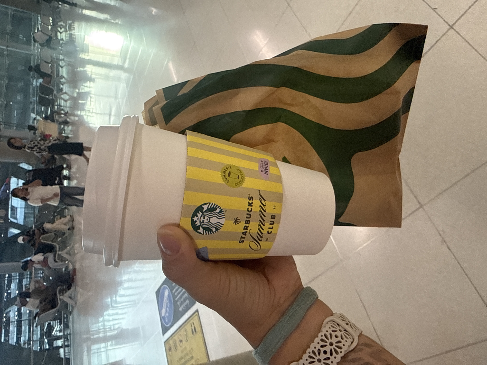A hand holds a white Starbucks coffee cup with a yellow and white striped sleeve that reads "STARBUCKS Summer CLUB". A brown paper Starbucks bag with green stripes is held behind the cup. The background shows a bright, tiled floor and rows of seating in what appears to be an airport or public waiting area. Several people are seated in the background, some looking at their phones. The lighting is bright and reflects off the polished floor. This scene is likely located in an airport terminal in Nong Prue Subdistrict, Thailand, given the presence of many travelers and airport-style seating.