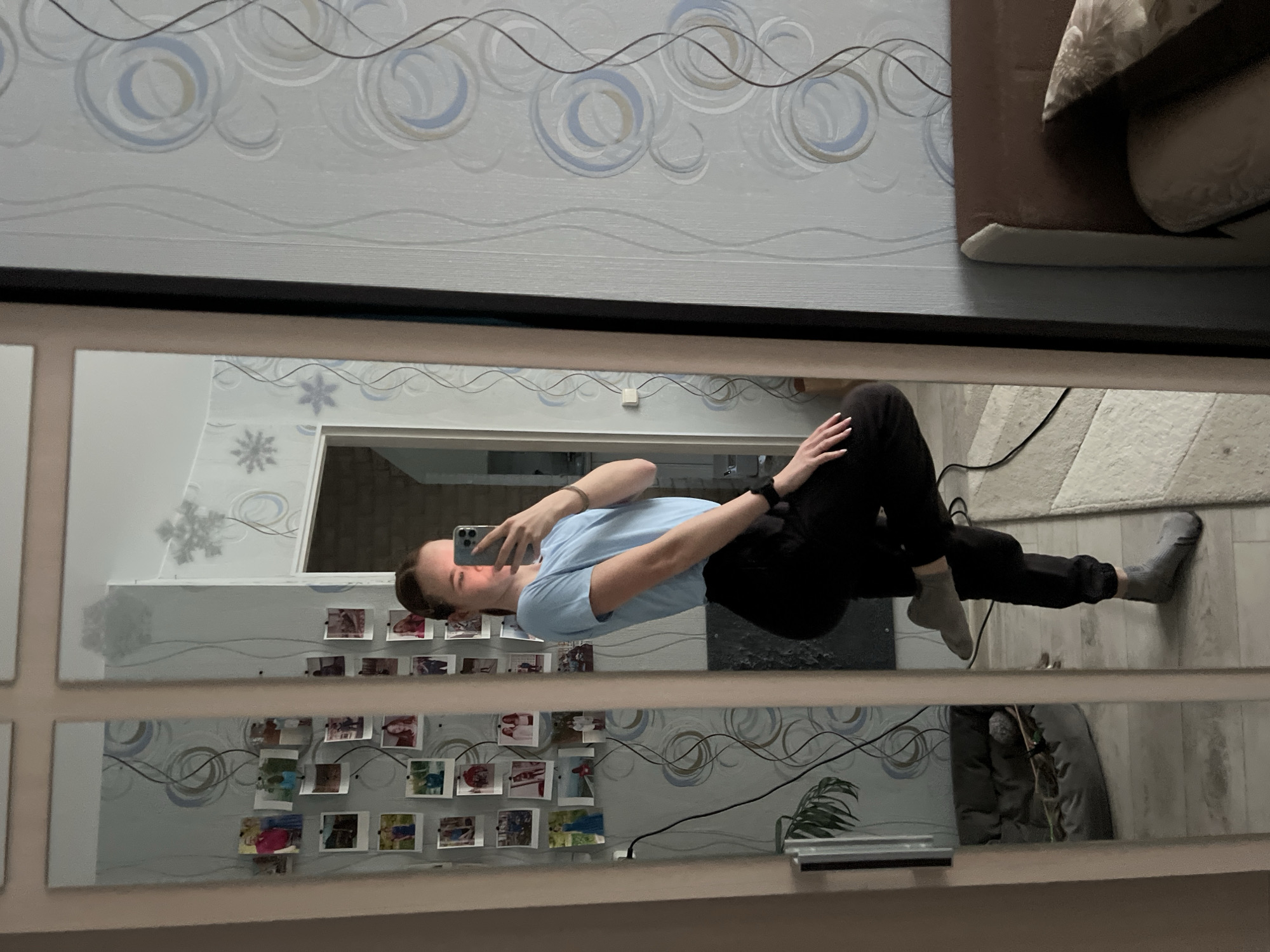 The image shows a reflection in a multi-pane mirror, seemingly taken in an apartment. The reflection features a person holding a phone and taking a selfie. The person is wearing a light blue t-shirt and black pants. The floor appears to be a light-colored laminate. A gray comforter or cushion rests on the floor. In the background, the wallpaper has a swirly pattern, and there are small photographs attached to the mirror. The presence of the Russian-style wallpaper design, along with the image of an interior apartment and objects such as houseplants, indicates a domestic setting in Russia. Based on the context of the request, it is likely located in the city of Chaikovsky.