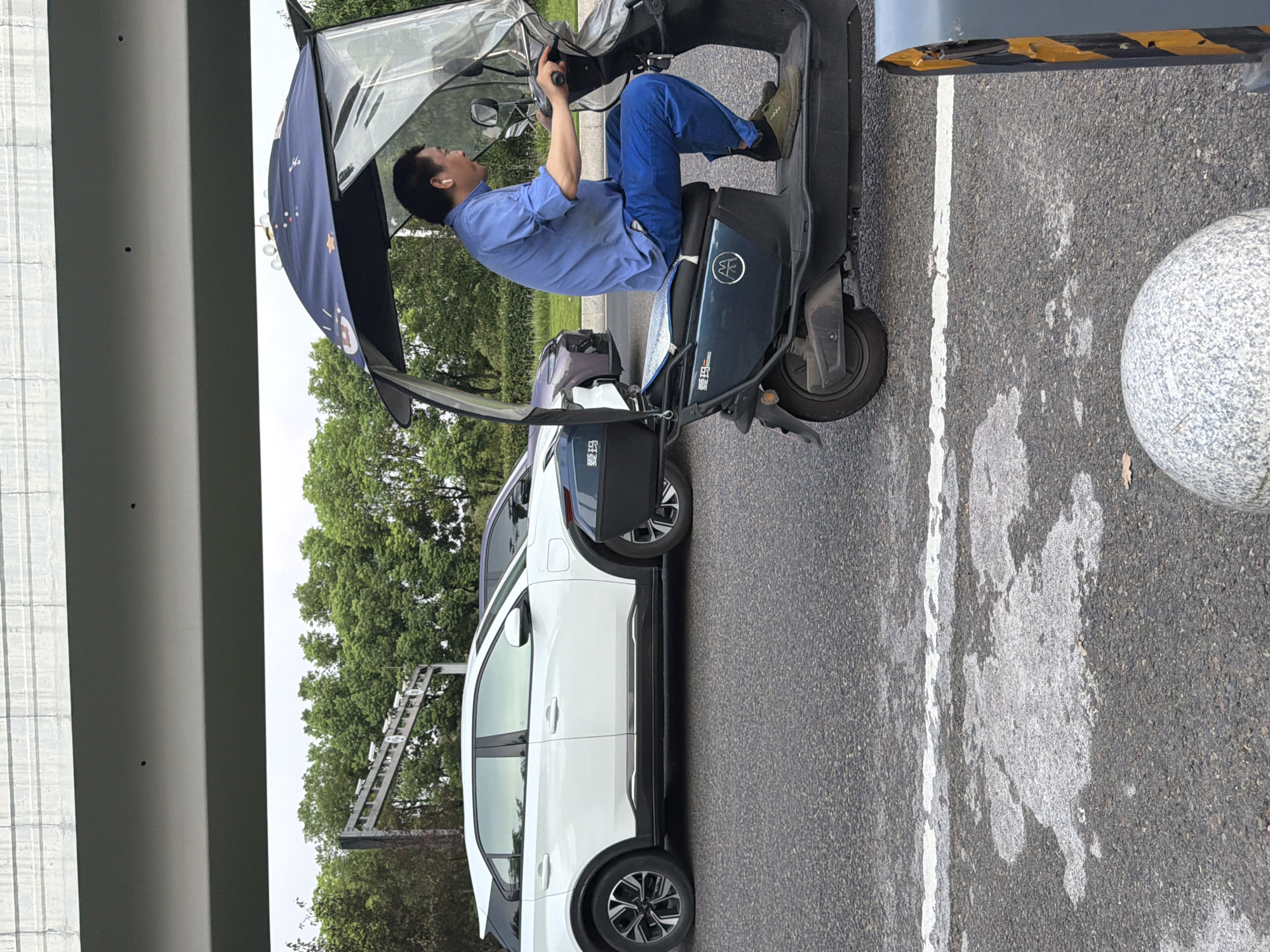 A man wearing a blue shirt and blue pants is riding a dark green three-wheeled electric scooter with a blue patterned canopy. He has white earbuds in his ears and is holding the handlebars of the scooter. The scooter has a silver emblem on its side. In the background, a white car is parked on the left, and a dark purple car is behind it. Trees and a bridge-like structure with cameras are visible behind the cars. The scene is on a paved road with a white line. The image appears to be taken in Jiangjin, China.