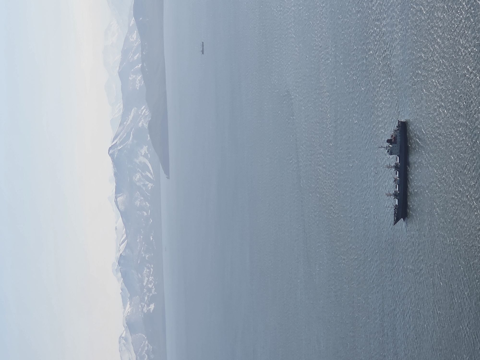An aerial, high-angle shot captures a wide expanse of calm, gray-blue ocean under a hazy sky. In the distance, a range of snow-capped mountains forms a hazy, blue silhouette against the pale sky. In the mid-ground, a small ship is visible on the water. In the foreground, a larger, dark-colored cargo ship is positioned slightly to the right, its deck structures clearly defined. The water exhibits gentle ripples, catching the light in a way that suggests a bright, though overcast, day. The scene depicts a serene maritime landscape with a backdrop of formidable mountains, likely in a remote, coastal region of Russia, possibly near Крутобереговый based on the context provided.