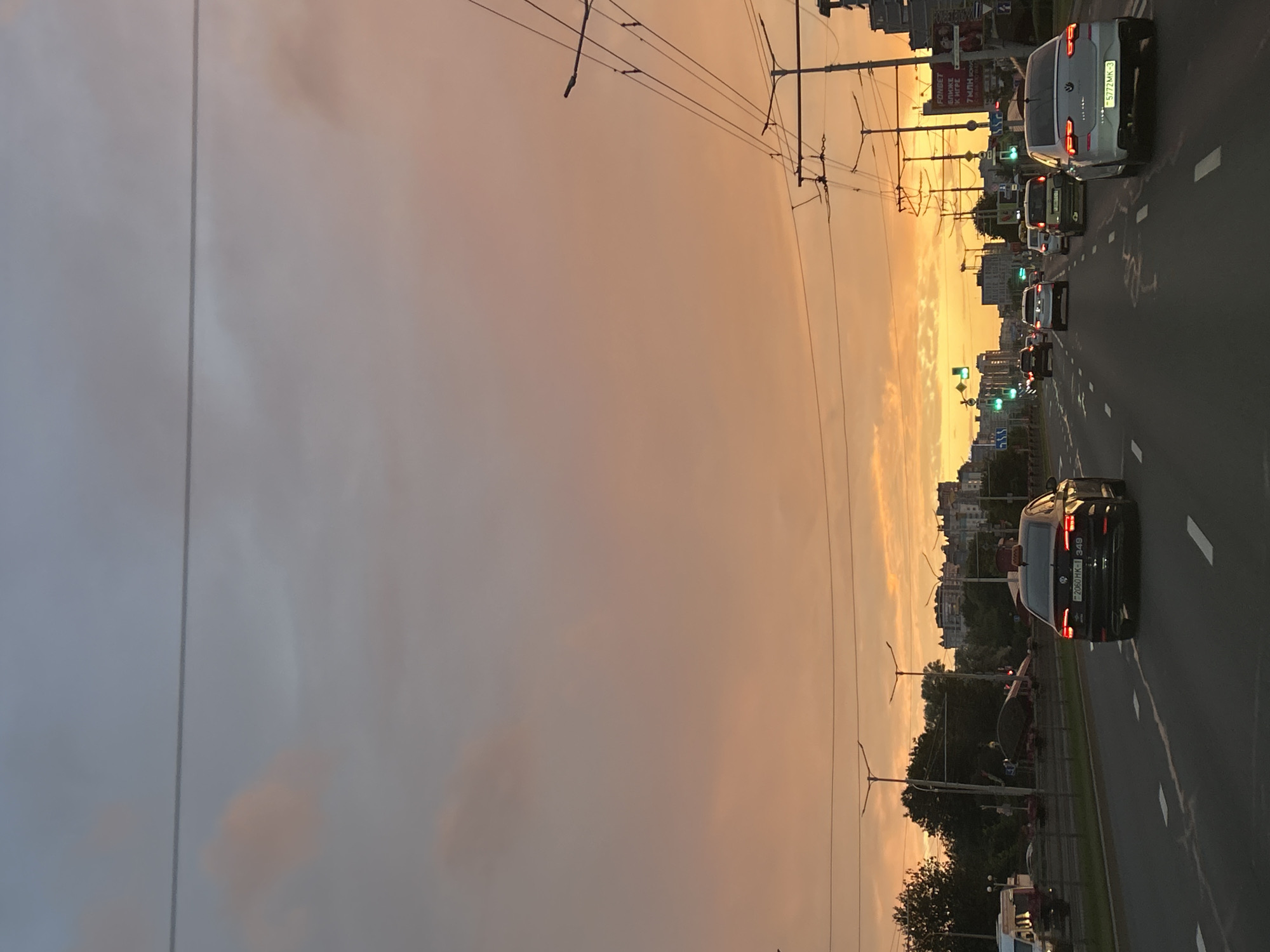 The image captures a street scene in Minsk, Belarus, during a sunset. The sky is a dramatic display of orange and pink hues, with scattered clouds. A multi-lane road runs through the scene, with several cars driving in both directions. Streetlights and overhead power lines are visible, indicating an urban environment. Buildings line the right side of the road, some of them appearing to be apartment complexes. The overall atmosphere is calm, with the setting sun casting a warm glow over the city.