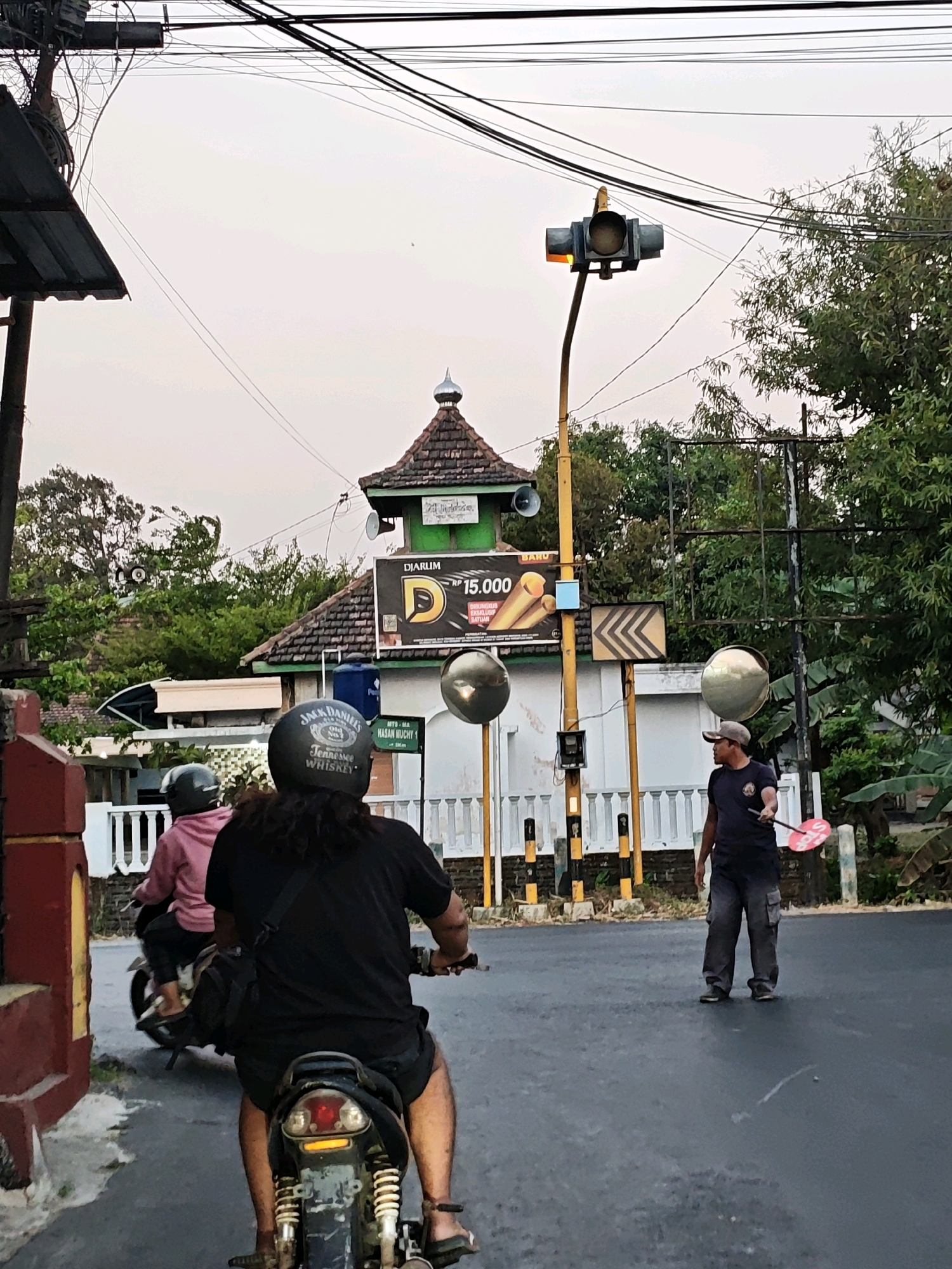 In Pagu, Indonesia, an outdoor scene depicts a road with two motorcycles and a pedestrian. The leading motorcycle, ridden by a person with long hair wearing a black helmet and t-shirt, is partially visible from behind. A second motorcycle, ridden by a person in a pink jacket and black helmet, is ahead of the first. A man, wearing a cap, t-shirt, and pants, stands on the side of the road holding a red stop sign. A traffic light with an amber light illuminated is visible above, accompanied by power lines and trees. A building with a brown roof and green accents, possibly a mosque or similar structure, is in the background, with an advertisement for "DJARUM" featuring a D logo and "Rp 15.000". The overall environment suggests a street scene in a populated area.