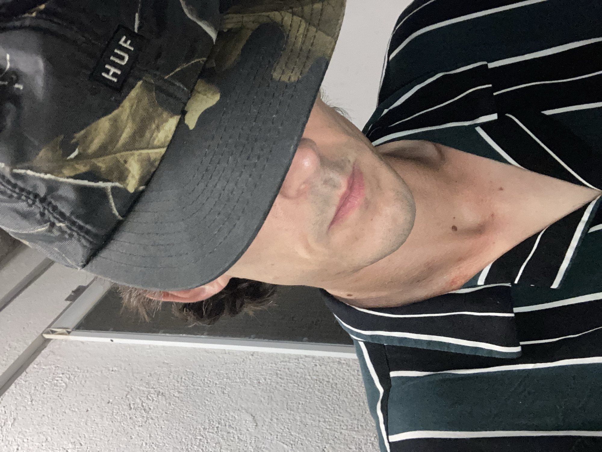 The image shows a close-up of a person wearing a camouflage patterned hat with a "HUF" logo and a dark green and white striped shirt. The hat is pulled down, obscuring the upper part of the face. The person is looking downwards and slightly to the left. The background is a textured wall and a doorway or window frame. There are no identifiable landmarks or features that would indicate a specific location within Sabadell, Spain.
