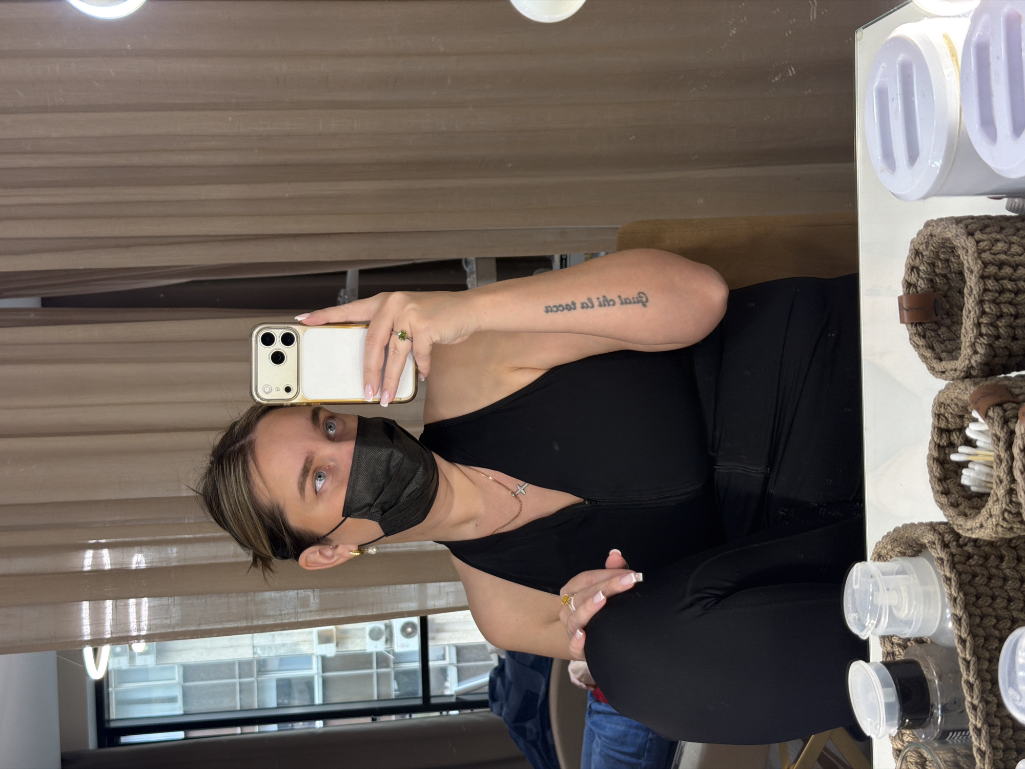 A woman is captured taking a mirror selfie in an indoor setting, likely a dressing room or vanity area, in Moscow, Russia. She holds a white and gold iPhone with a clear case and triple camera array, reflecting a ring light. She wears a black face mask, partially obscuring her face, but her blue-grey eyes are visible. Her hair is styled with a side part, showing brown strands with lighter highlights.
She is dressed in a black top or dress, and accessorized with a gold necklace featuring a small cross pendant. On her left ring finger, she wears a ring with a green stone, and on her right ring finger, another with a yellow/amber stone. Small gold hoop earrings with dangling pearls are also visible. A tattoo, appearing reversed in the mirror, is visible on her right forearm, which reads "quál chi la tocca" (Italian for "Whoever touches it").
The background features beige/brown horizontal blinds or curtains. Through a window behind her, an urban landscape with buildings and an air conditioning unit can be seen, suggesting daytime. To the right, a white shelving unit holds brown crocheted baskets, containing various personal care items like cotton swabs and clear dispenser bottles. The overall scene suggests a calm, personal moment in a well-lit interior space.