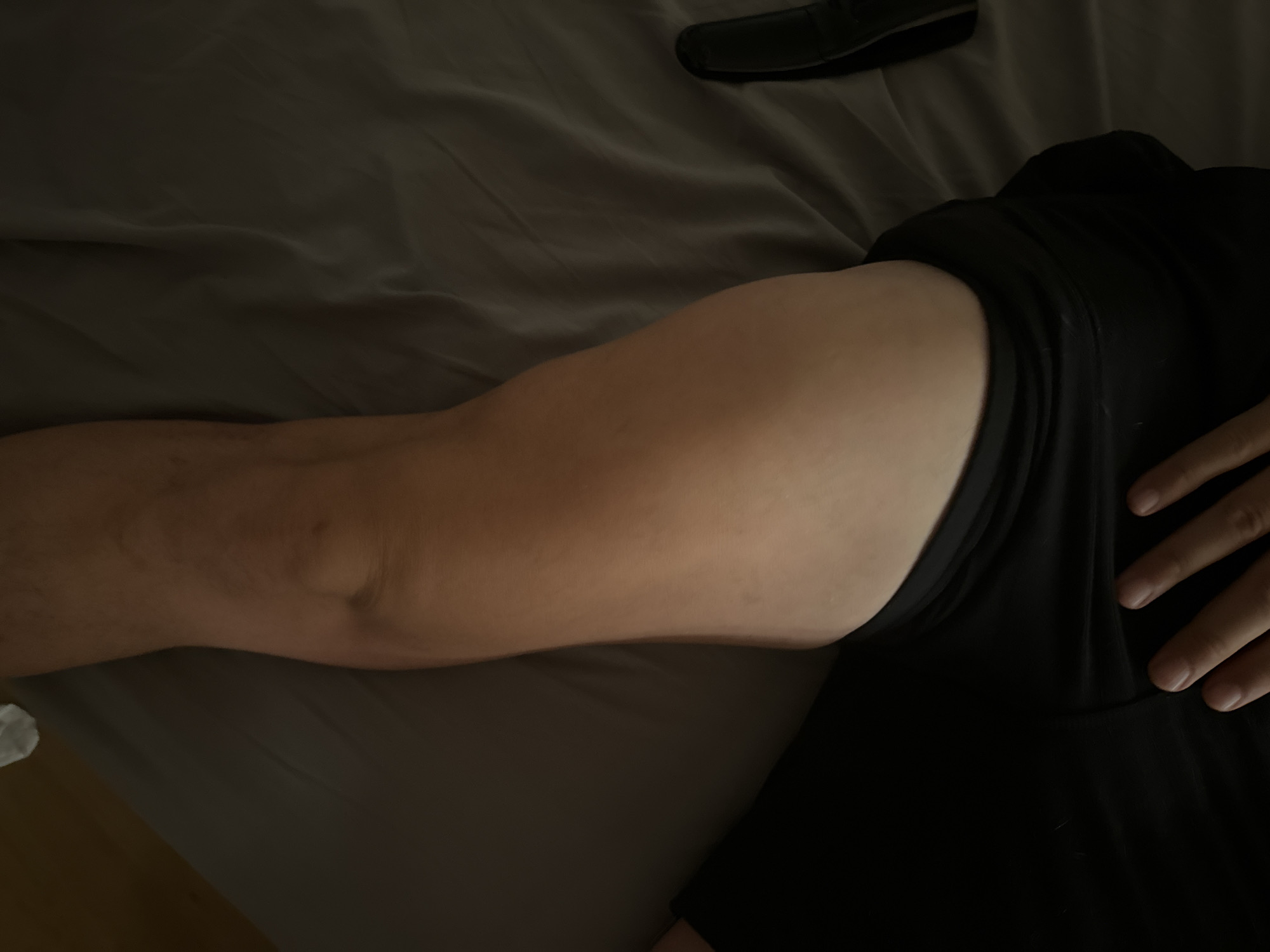 The image shows a close-up view of a person's thigh and part of their torso, clad in black shorts. The person's skin is fair. The scene appears to be indoors, possibly on a bed, indicated by the presence of a gray wrinkled sheet. The lighting is dim, casting shadows across the scene. A black object, resembling a shoe or bag, is visible on the right side of the frame. There is no discernible context to place this image within the city of Murcia, Spain.