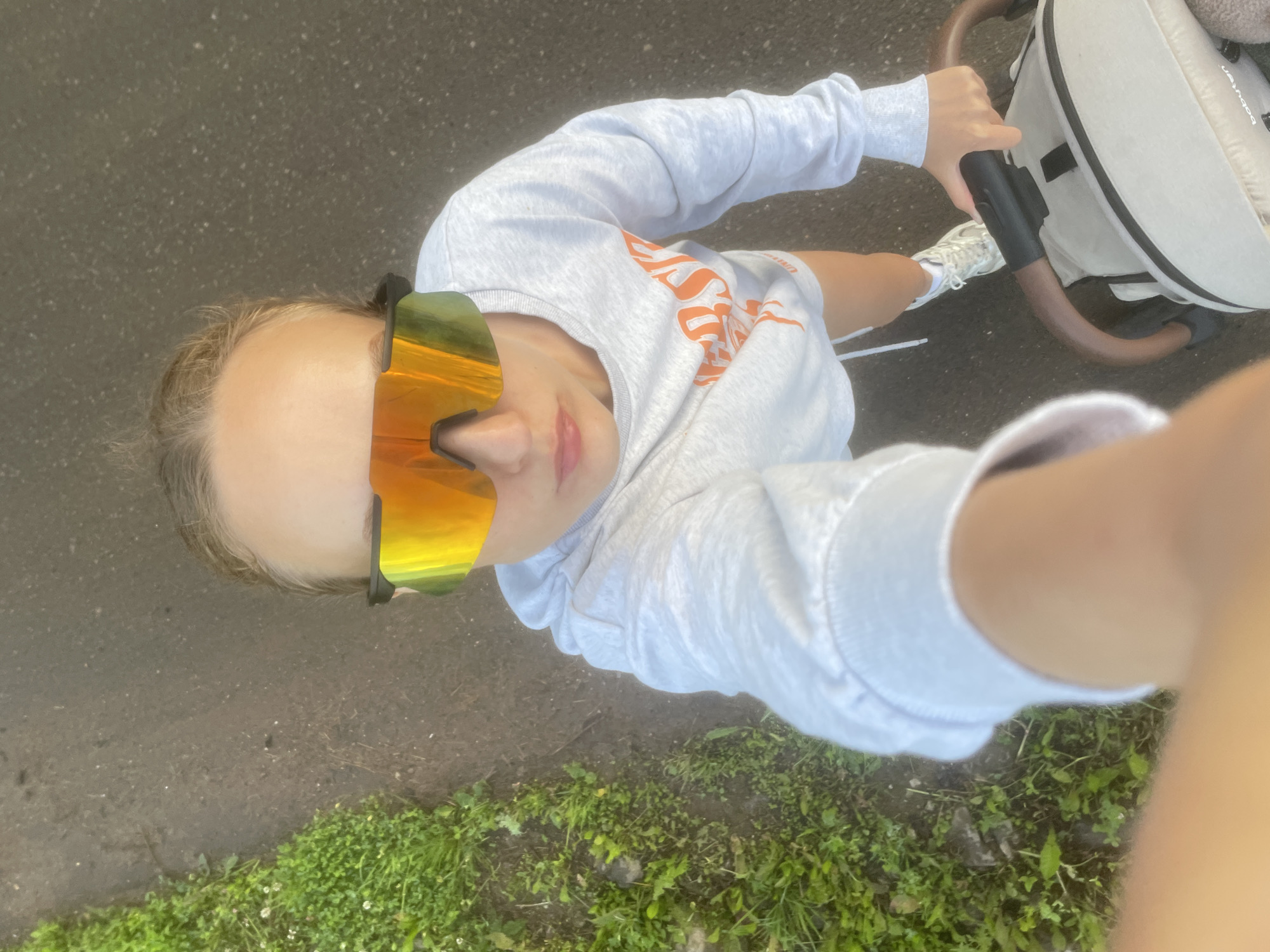 A young woman with blonde hair pulled back is taking a selfie while pushing a stroller. She is wearing a light grey sweatshirt with orange lettering and white athletic shoes. The sweatshirt has the word "VERSACE" printed on it. She is also wearing mirrored wrap-around sunglasses with orange and green lenses. The stroller is light-colored with a black and brown handle. The woman is standing on a paved path with green grass and clover on the left side. The background is a dark, possibly wet, paved surface. The lighting suggests it is daytime. The setting appears to be an outdoor area, possibly a park or a residential street, in the city of Podshivalovo, Russia.