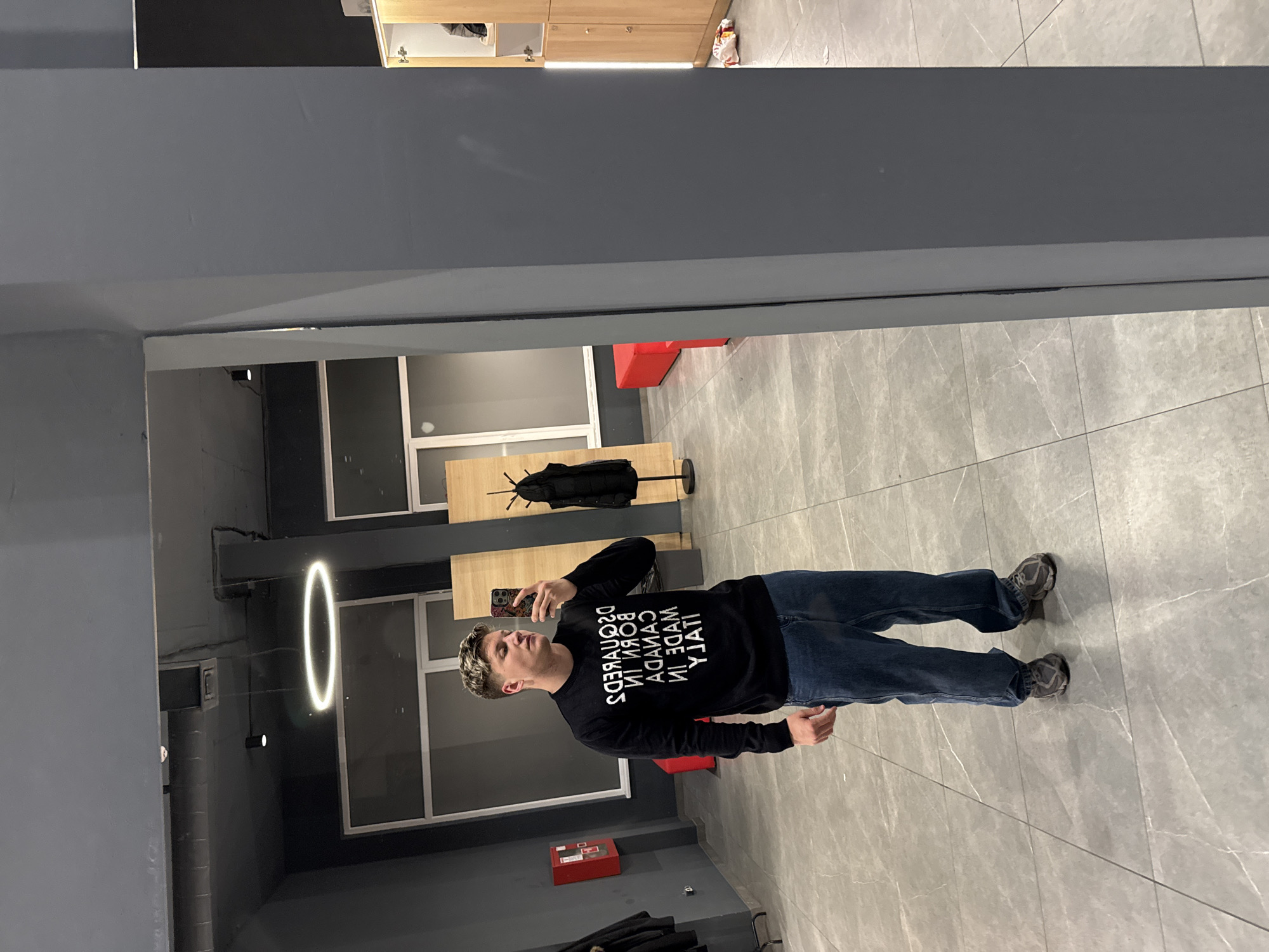 A full-length photograph shows a man standing in what appears to be an interior space, likely a store or business. The man is dressed in jeans and a black sweatshirt with large, stylized white text on the front. He is holding a mobile phone up as if taking a selfie, looking toward the device. He seems to have light blonde hair.
The environment features light grey-colored tile flooring, and the walls are a combination of light grey and dark grey/black. In the background, there's a circular light fixture, and a coat rack is situated near the wall. There are also visible windows with white frames.
Without specific landmarks or building identifiers, it's impossible to definitively locate this scene in Plovdiv, Bulgaria, from the details of this single photograph alone.