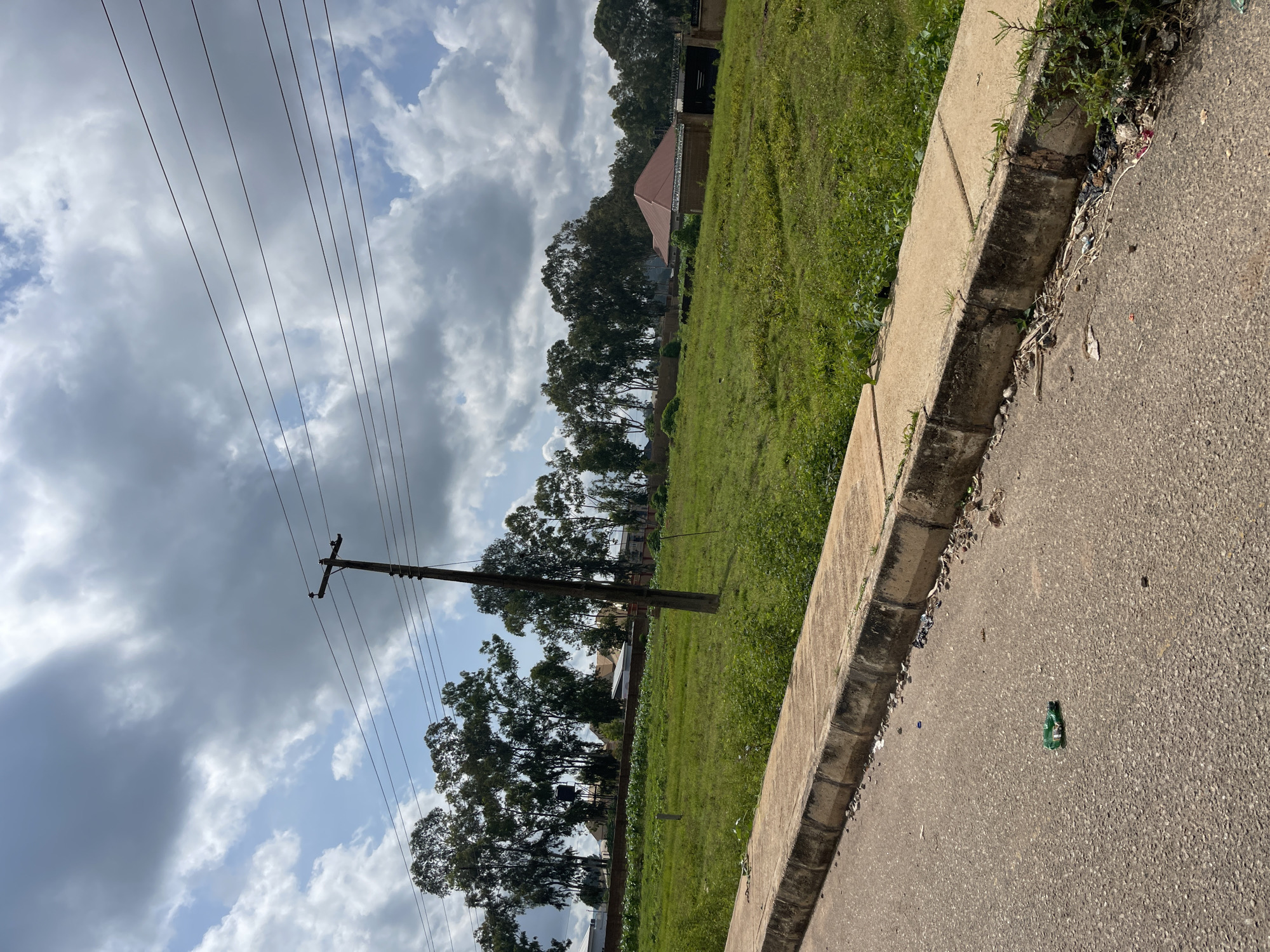 The image shows a street scene in Jos, Nigeria, featuring a paved road in the foreground with a concrete curb and a sidewalk. A discarded green plastic bottle lies on the road. Beyond the sidewalk is a large, grassy field with some scattered vegetation. In the middle ground, a wooden utility pole with power lines stretches across the sky. Behind the field, a line of tall trees forms a natural barrier, with several buildings visible through them, including a house with a reddish-brown roof. The sky is partly cloudy with a mix of gray and white clouds. The overall impression is one of an undeveloped or developing area on the outskirts of a city.