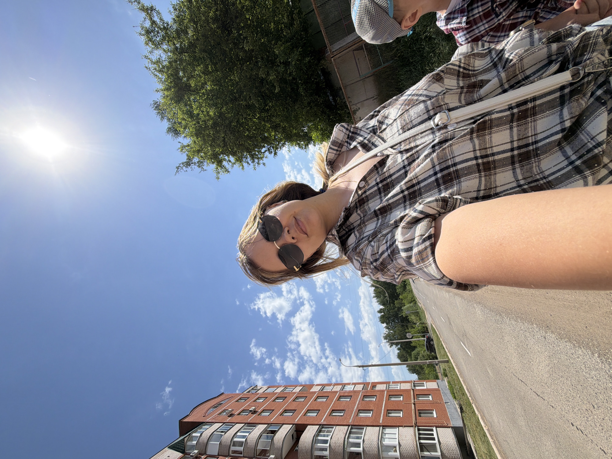 A woman wearing a plaid shirt and sunglasses takes a selfie outdoors on a sunny day, with a red brick apartment building with white-framed windows visible in the background. A child wearing a plaid shirt and baseball cap is partially visible on the right side of the frame. A paved road runs parallel to the building. Trees and a blue sky with some clouds are visible in the background. The location is Chaikovsky, Russia.