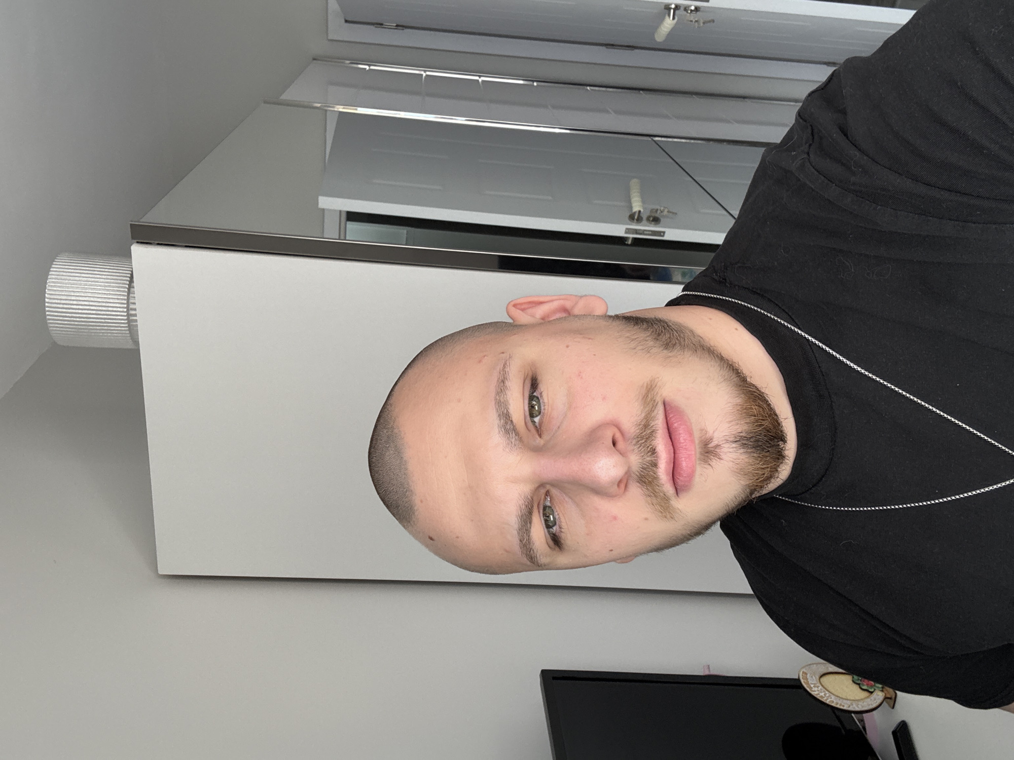 A man is posing for a photo indoors. He has a shaved head and a short beard and is wearing a black t-shirt and a silver chain. Behind him is a white cabinet, possibly in a kitchen or bathroom, with a chrome edge. A white louvered door is visible above the cabinet. A black television is partially visible in the lower portion of the frame. The location is not discernible from the image.