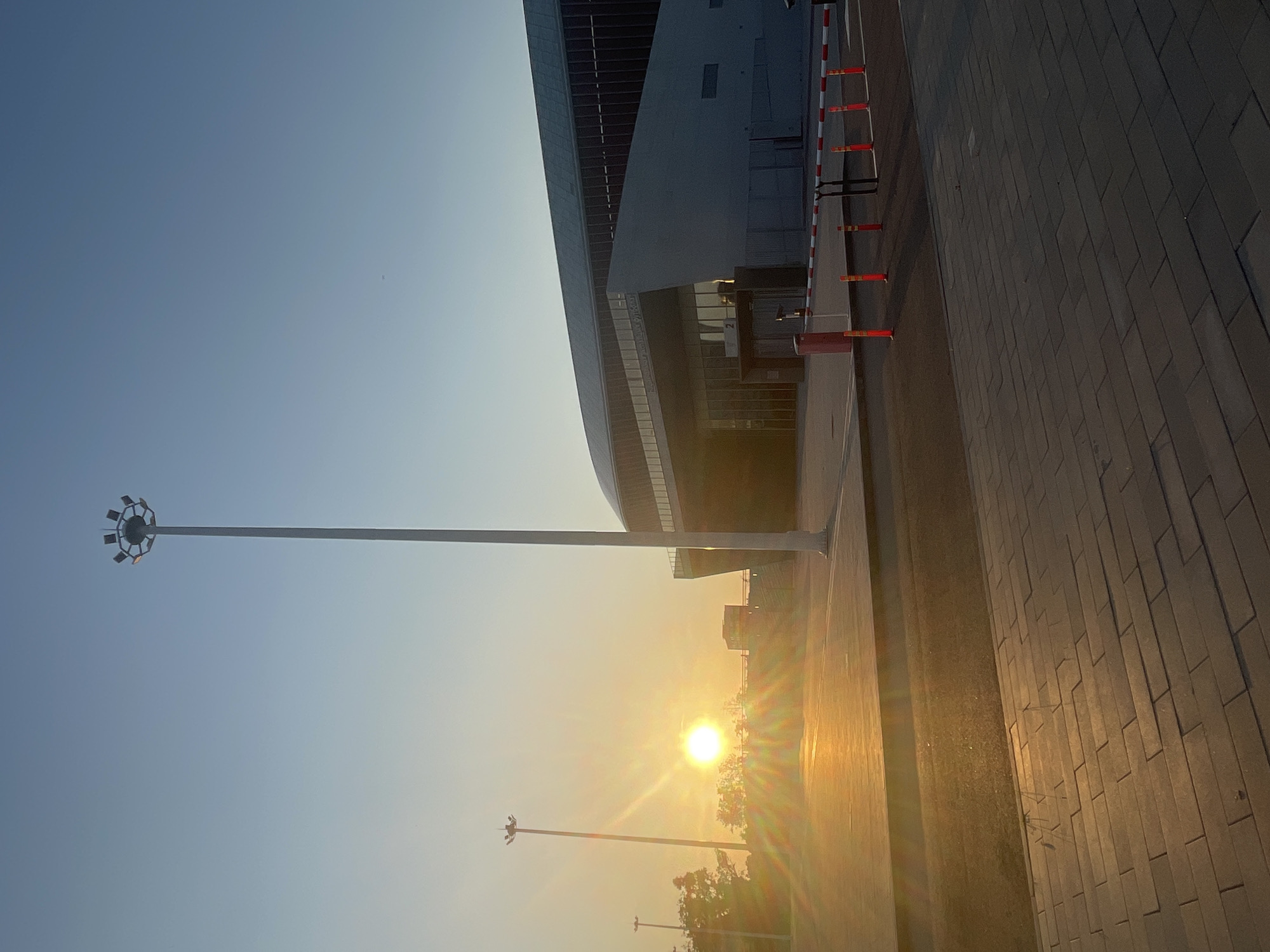 The image depicts a modern, somewhat curved building with a dark facade and large glass windows. The building appears to be a stadium or a large public venue. In the foreground, a paved walkway with a brick-like pattern runs alongside the building. A tall lamppost with multiple lights is visible on the left side of the frame.
The sun is setting, casting a warm, golden light across the scene and creating lens flares. The sky is a clear, gradient blue, transitioning to a pale yellow near the horizon. The low angle of the sun suggests it is late afternoon or early evening. There are some temporary barriers with red and white stripes set up along the edge of the walkway, possibly for crowd control or construction. In the distance, more trees and smaller buildings are visible, silhouetted against the bright sunset. The overall mood is peaceful and serene, enhanced by the warm lighting. There is no visible text in the image.