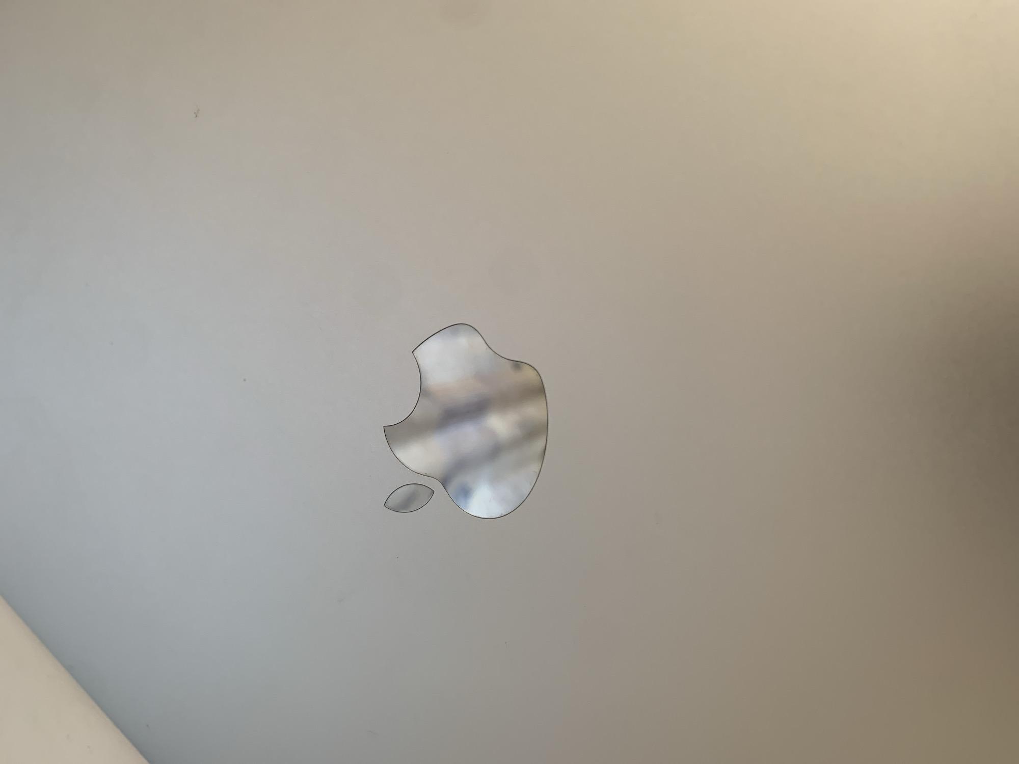 The image is a close-up, overhead shot of the Apple logo on the lid of a silver laptop. The logo is metallic and reflects the surroundings, showing some blurry light and color variations within its shape. The laptop's surface appears smooth and matte, with some faint smudges or dust specks visible. The lighting suggests an indoor environment, likely from a window or overhead light source, casting a warm glow on the right side of the image. There are no people or other objects in the frame, and no identifiable landmarks that would indicate a specific location in Октябрьский, Russia. The scene is static and focuses solely on the detail of the Apple logo.
