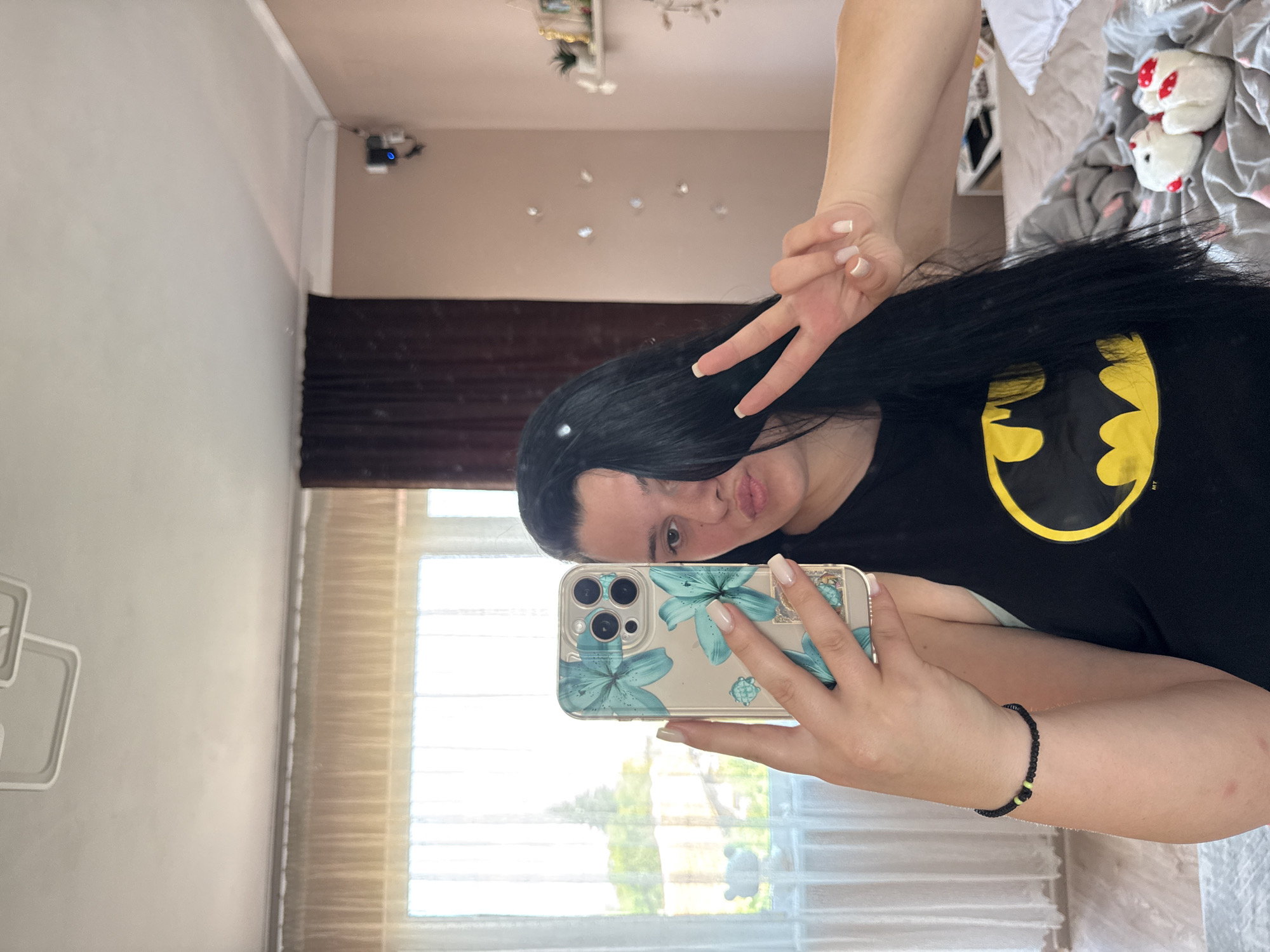 This mirror selfie, taken indoors in Rogosh, Bulgaria, features a young woman posing playfully during what appears to be daytime. She has long, straight dark hair and is wearing a black t-shirt adorned with a prominent yellow Batman logo, alongside "TM" text. In her right hand, she holds a smartphone with a clear case decorated with teal/blue floral patterns and a small sea turtle. Her left hand is raised in a peace sign, and her lips are slightly puckered. Her fingernails are painted a light, neutral color. A black beaded bracelet with two small yellow/lime green beads is visible on her right wrist.
The setting appears to be a private room, possibly a bedroom or living area. Reflected in the mirror is a bed with a gray blanket featuring small pink hearts, on which two white, chicken-like stuffed animals with red accents are resting. A white geometric wall decoration is mounted on the wall to the left. Several small, circular light reflections or decorative elements are scattered on the wall above the bed. Sunlight streams through a large window, visible in the reflection, which has sheer white curtains and what appears to be venetian blinds. Green trees and foliage are visible outside the window. A dark brown curtain is seen hanging in the actual room. Near the ceiling, a small, dark device with a blue indicator light is mounted on the wall. The overall mood is casual and relaxed.