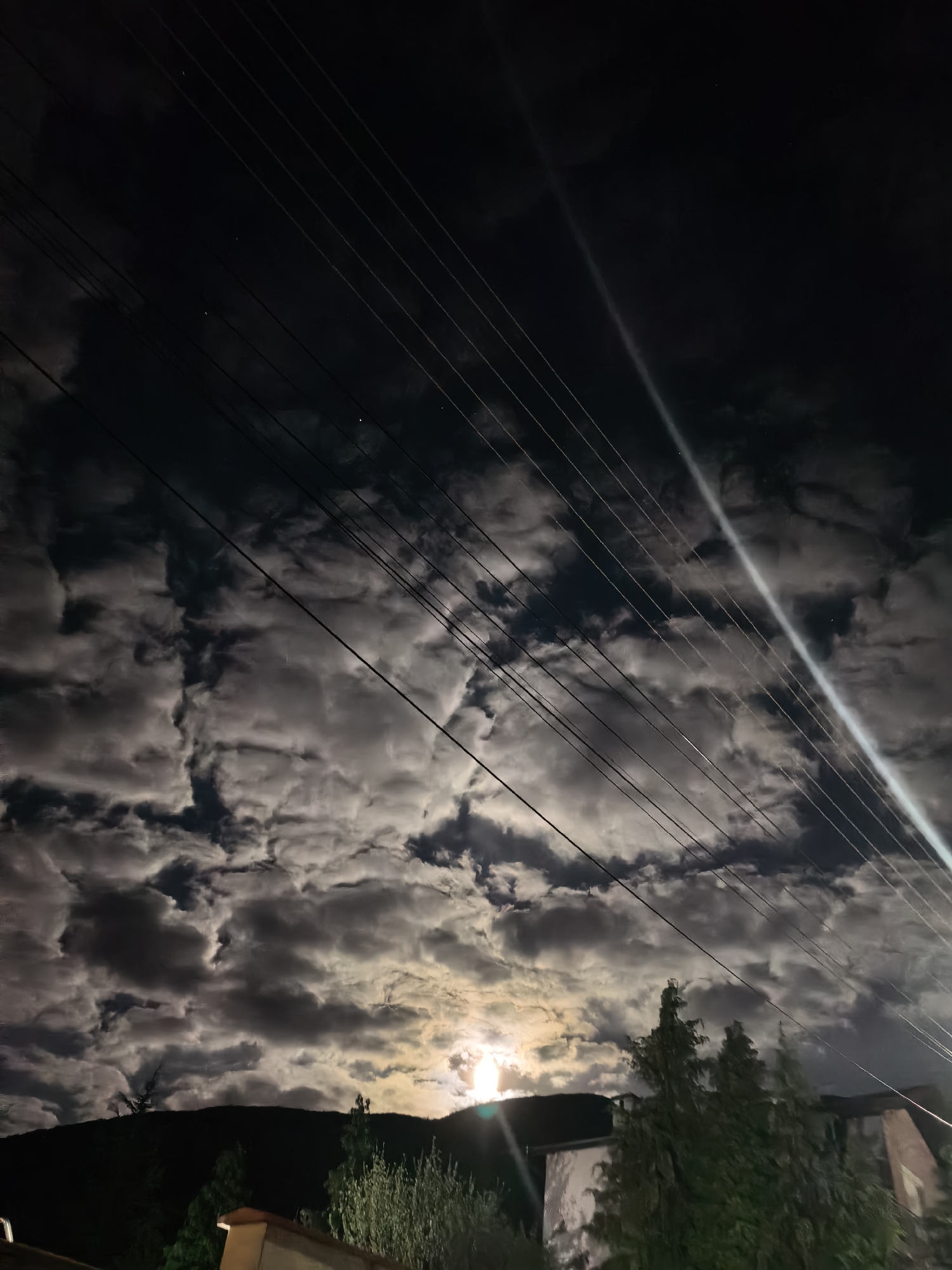 The image displays a dramatic night sky over Sapareva Banya, Bulgaria, dominated by dense, illuminated clouds. The sky is dark, but the clouds glow brightly from behind, suggesting a strong light source, likely the moon, partially obscured and casting light through and around them. These clouds exhibit varied textures and patterns, with brighter, lighter areas contrasting with darker, shadowed sections.
Several thin, dark power lines or utility wires stretch diagonally across the upper and middle portions of the sky, adding a man-made linear element to the natural cloudscape. A prominent, bright, diagonal light streak or lens flare is visible, extending from the upper right towards the mid-left, likely originating from the intense moonlight. Faint stars can be observed in the clearer, darker patches of sky between the clouds.
Below the sky, the dark silhouettes of mountains or hills form the horizon line. In the foreground, parts of lush green trees and what appear to be the roofs and walls of buildings are visible, partially illuminated by the ambient light. The scene captures a still moment of a cloudy, moonlit night, creating an atmospheric and somewhat mysterious ambiance. No specific activity or people are depicted, and no text is visible.