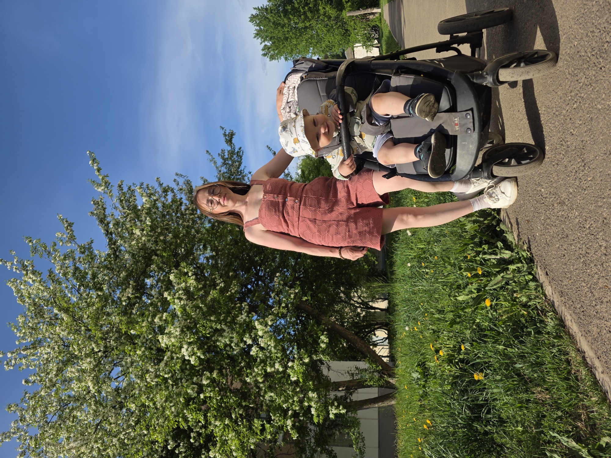 A woman stands with a baby in a stroller on a paved sidewalk in front of a grassy area and a tree with white blossoms under a blue sky. The woman is wearing a rose-colored dress and white sneakers. The baby in the stroller wears a patterned hat and jacket. The stroller is positioned on the sidewalk. Yellow flowers are visible in the grass. The location is Chaikovsky, Russia.