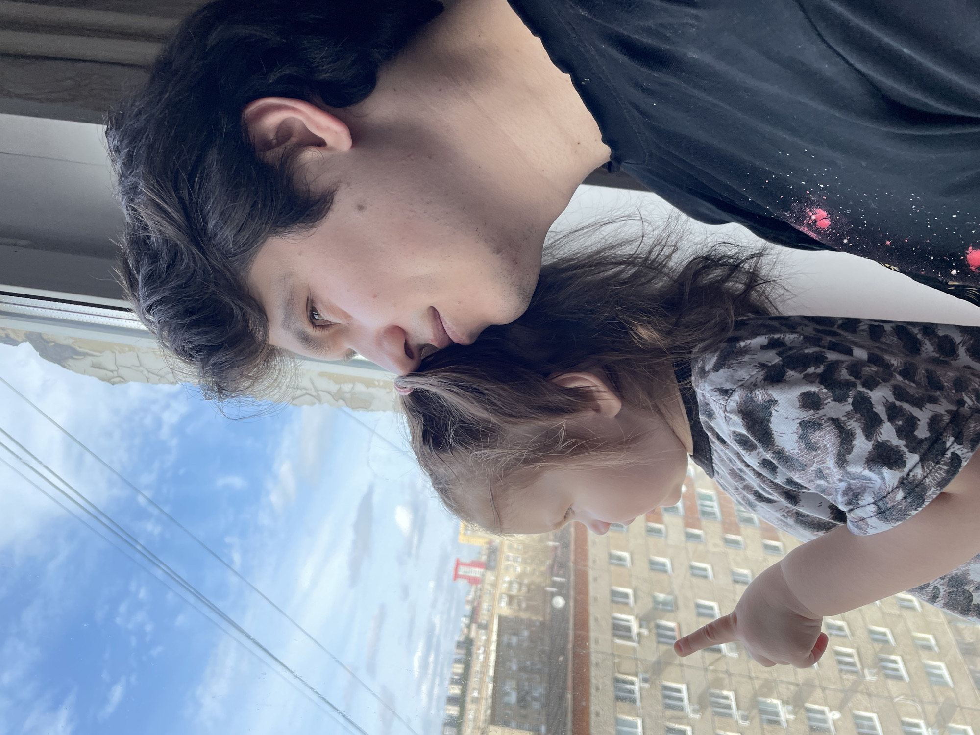 A medium, eye-level shot depicts a man and a child gazing out a window in Izhevsk, Russia. The man, fair-skinned with dark hair, wears a black t-shirt with splotches of red paint and rests his head near the child’s head. The child, with light brown hair pulled back with a pink hair tie, is dressed in a grey and black animal-print shirt. The child is pointing toward a light brown building with rows of windows outside the window. The sky visible through the window is a light blue with scattered white clouds.