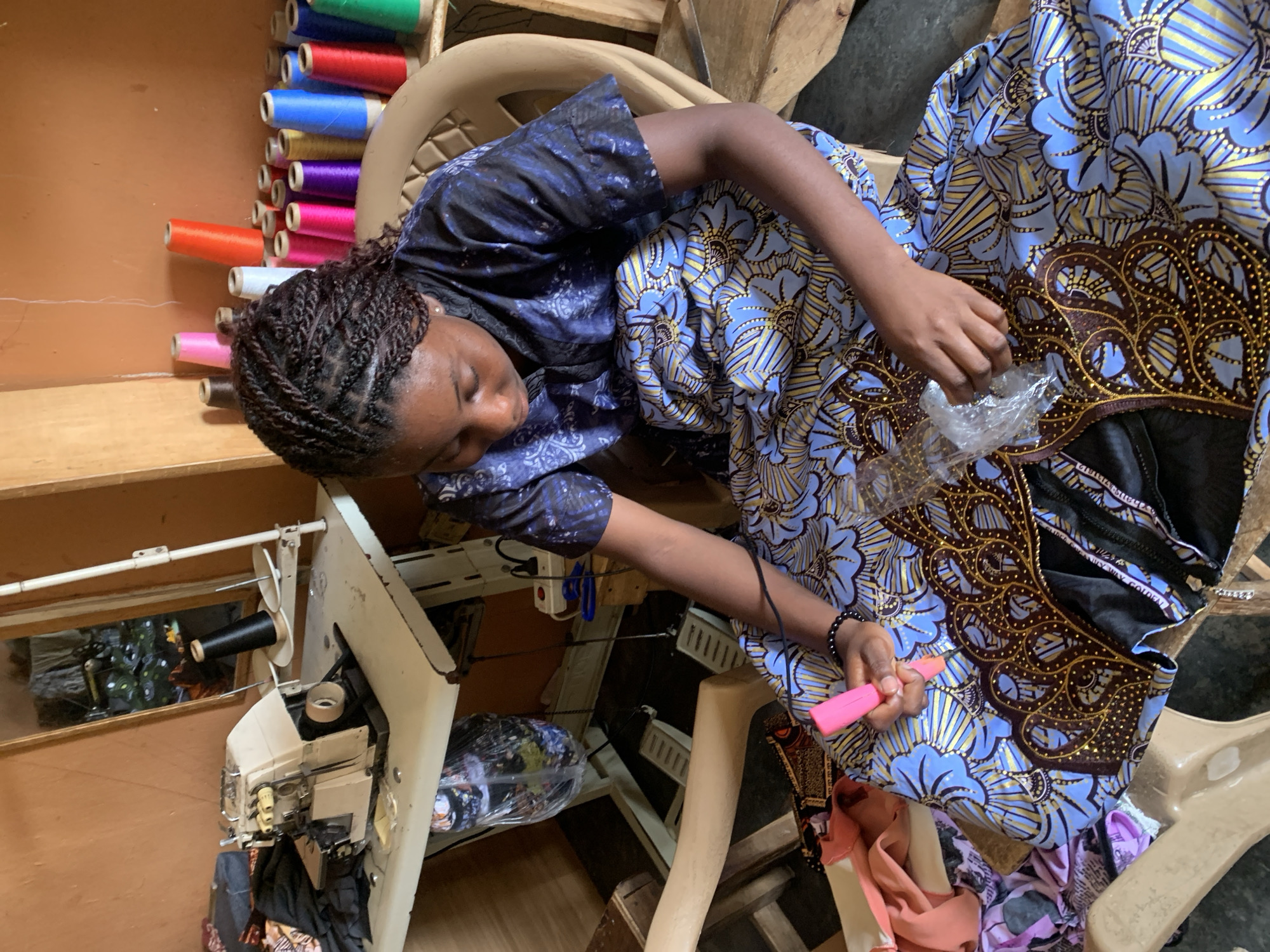 In a cluttered workshop in Jos, Nigeria, a young woman with braided hair sits on a beige plastic chair, engrossed in her craft. She wears a blue and black patterned dress and is hunched over a vibrant blue and gold fabric, adorned with intricate floral and crown-like embroidery. Her hands are busy, one holding a pink tool, possibly a seam ripper or stitch remover, while the other manipulates a piece of transparent plastic covering some of the embellishments.
The background reveals the operational chaos of a sewing studio. To her left, a vintage white sewing machine sits on a worn metal table. Several rolls of fabric, some bagged, are stacked nearby. Behind her, a wall displays an array of colorful thread spools in shades of pink, orange, purple, blue, and green, arranged in a haphazard fashion. A mirror on the wall reflects more of the workshop's contents, including a sewing machine and hanging garments. Underneath the table, a power strip with plugs is visible, suggesting the electrical setup for the machinery. Wooden chairs and more fabric scraps are scattered around the space, contributing to the impression of a busy, active workspace. The floor appears to be concrete.