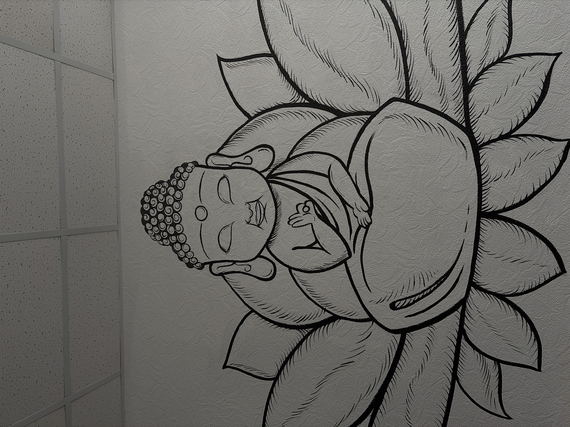 A grayscale image depicts a mural of a reclining Buddha in a lotus flower. The mural is located on a wall next to a suspended ceiling, possibly in an interior space. The mural is rendered in black outlines on a gray textured surface. The location is Chaikovsky, Russia.