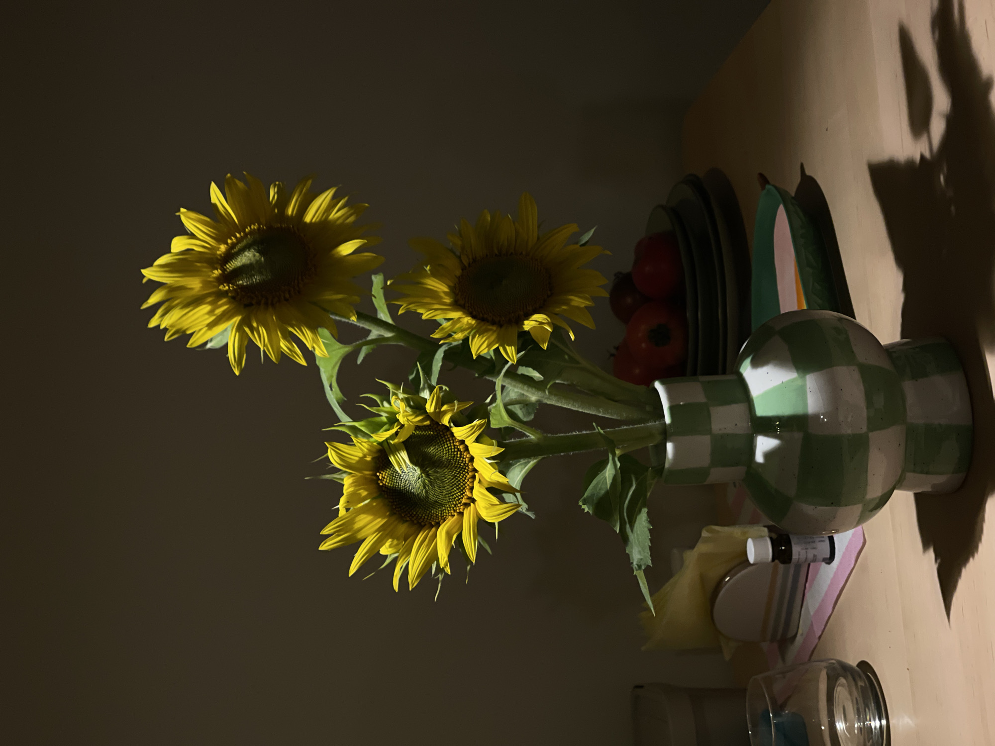 A still life photograph depicts three sunflowers in a green and white checkered vase. The sunflowers are positioned on a light-colored wooden surface, with the vase in the foreground. Behind the vase, a stack of dark green plates is visible, topped with a few red tomatoes. To the left of the vase, a clear glass is present, along with a small dark brown bottle and a folded yellow napkin. The background is dark, with a concentrated light source illuminating the sunflowers and casting shadows on the table. The scene is indoors, likely in a domestic setting, as suggested by the objects present. The photograph is taken from a slightly low angle, emphasizing the sunflowers.