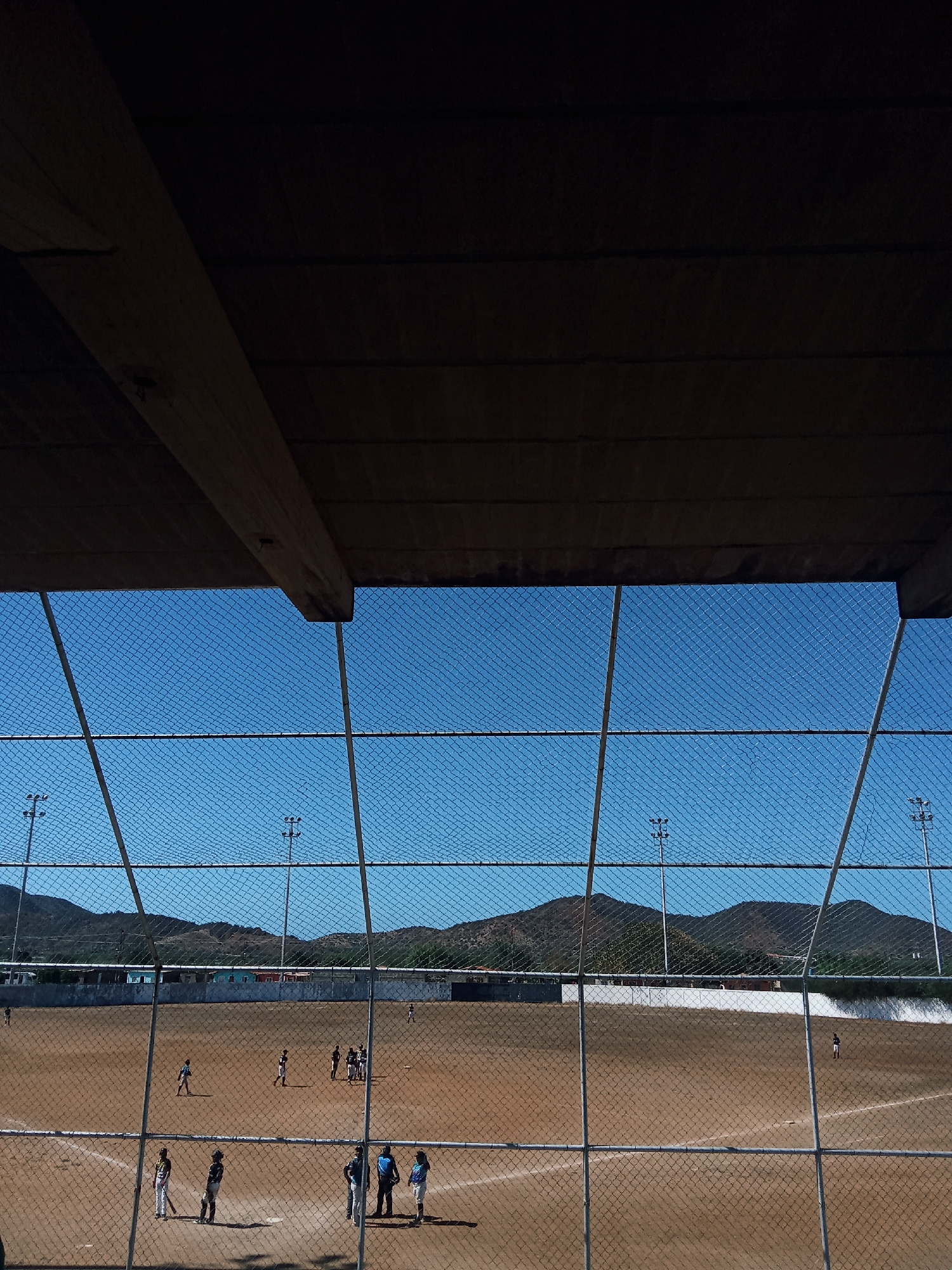 A baseball field is viewed through a chain-link fence, with a dark, wooden roof structure partially obscuring the top of the frame. The field is mostly dirt, with base lines marked in white. Several people, likely baseball players, are dispersed across the field. Beyond the outfield fence, there's a low white wall, followed by a line of buildings. In the background, hills or small mountains rise against a clear blue sky. The location is likely Juan Griego, Venezuela. Light poles are visible around the field.