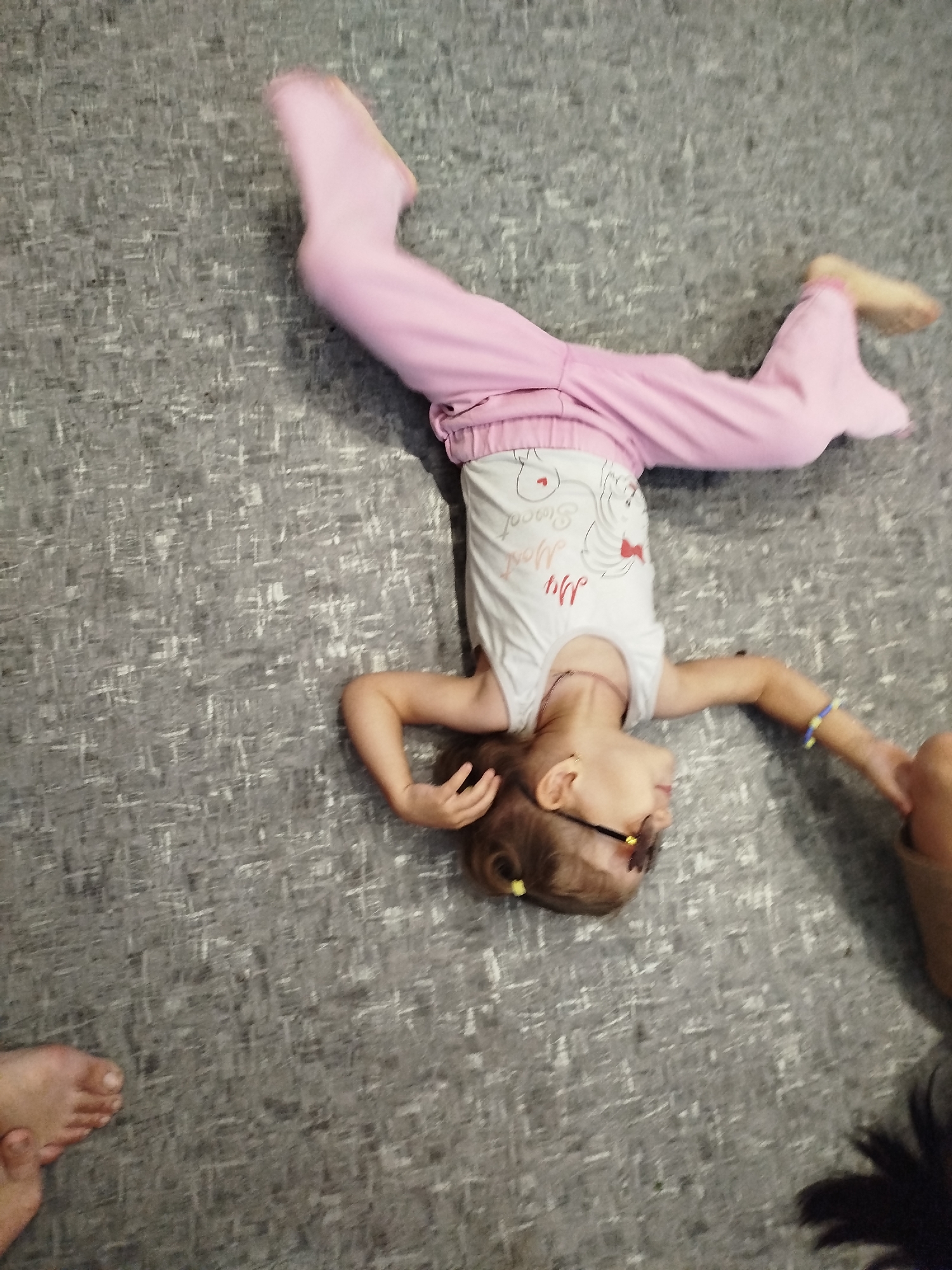 The media file shows a young girl performing a handstand on a grey carpeted floor. She is wearing pink leggings and a white t-shirt with a graphic design. She has a necklace and sunglasses on. A child's foot is visible in the bottom left corner of the frame, and a portion of an adult's hand is visible in the top right. The setting appears to be indoors, possibly a home, in Krasny Gulyai, Russia. The lighting suggests daytime. The overall impression is playful and active. The t-shirt has the text "My Sweet Home" printed on it.