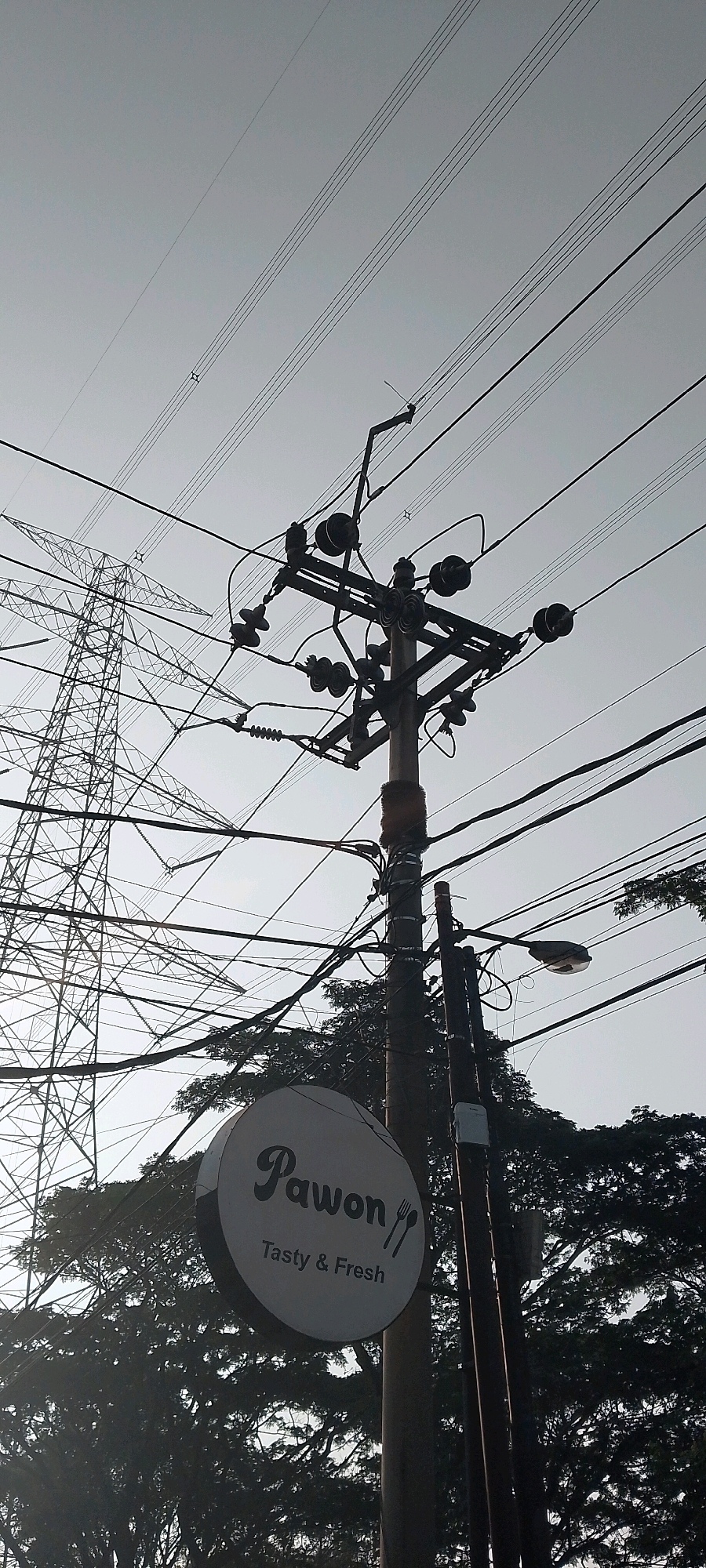 The image depicts an outdoor scene dominated by electrical infrastructure against a bright, overcast sky. In the foreground, a utility pole stands prominently, adorned with numerous wires, insulators, and what appears to be a circular sign. The sign is white with a dark border and features the word "Pawon" in a stylized font, followed by "Tasty & Fresh" and an icon of a fork and knife. To the left of the pole, a large transmission tower, also made of metal lattice, is visible. Trees with dense foliage are partially obscured behind the electrical equipment and the sign. A street lamp is attached to a secondary pole situated to the right of the main utility pole. The overall impression is one of urban infrastructure intertwined with natural elements. Based on its content and visual characteristics, this image is likely from Satriamekar, Indonesia.