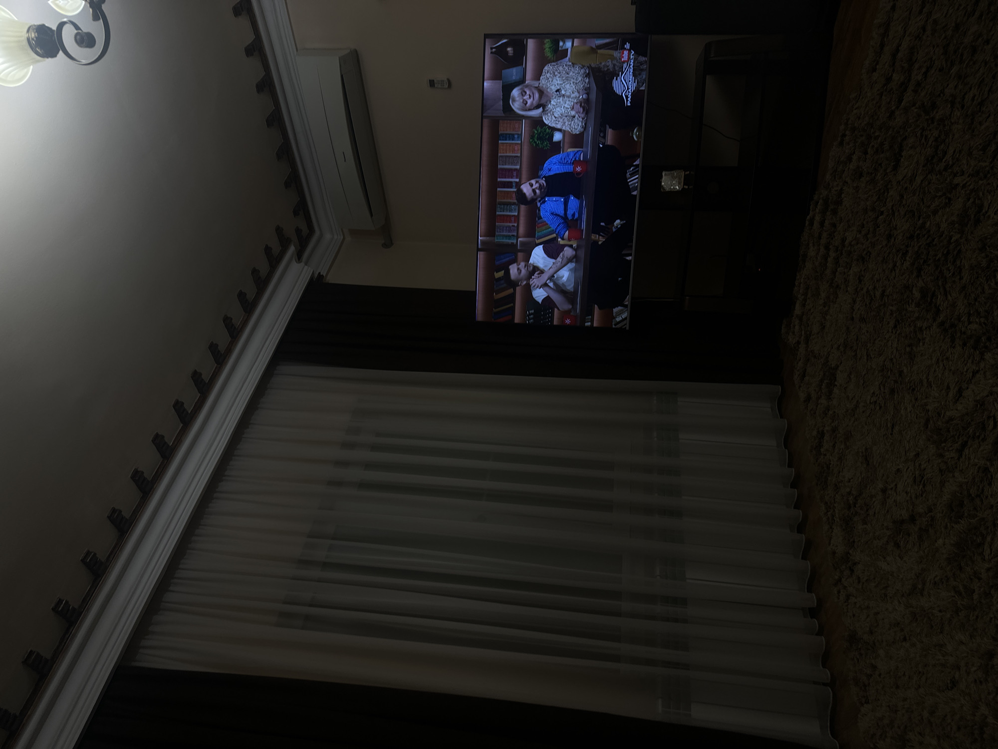 A dimly lit room features a television displaying an image of three individuals seated at a table, possibly engaged in a discussion or interview. The room has curtains drawn across a window and a visible ceiling fixture, along with a dark-colored rug. An air conditioning unit is mounted high on the wall. The city of Yerevan, Armenia is not verifiable from the image.
